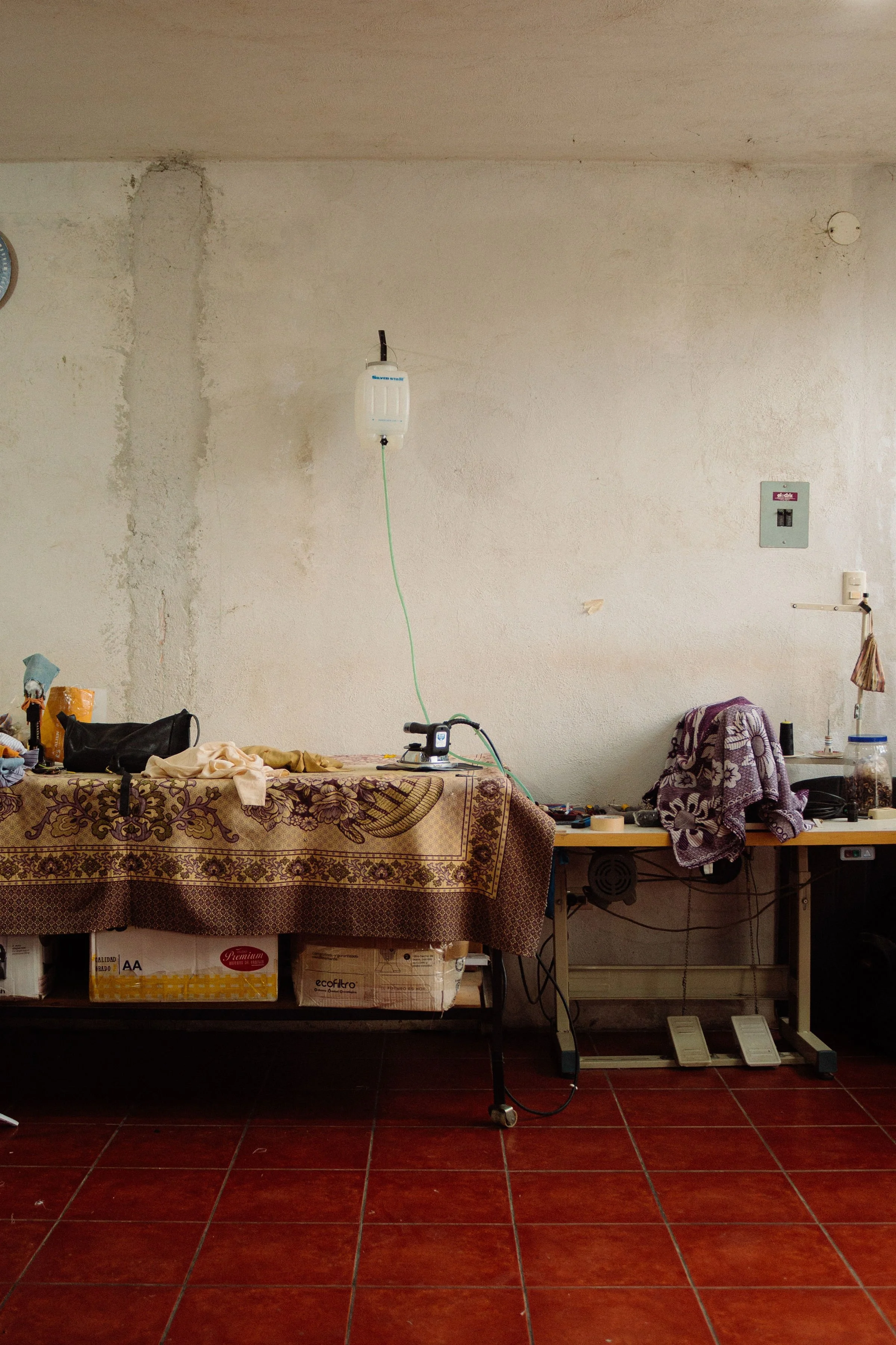 A cluttered workshop or storage area with a table covered by a patterned cloth, various objects, and a machine with a green cable hanging from the ceiling. The background consists of a wall with visible mold or water damage, a switchboard, and a small table with a cloth and items. The floor is made of red tiles.