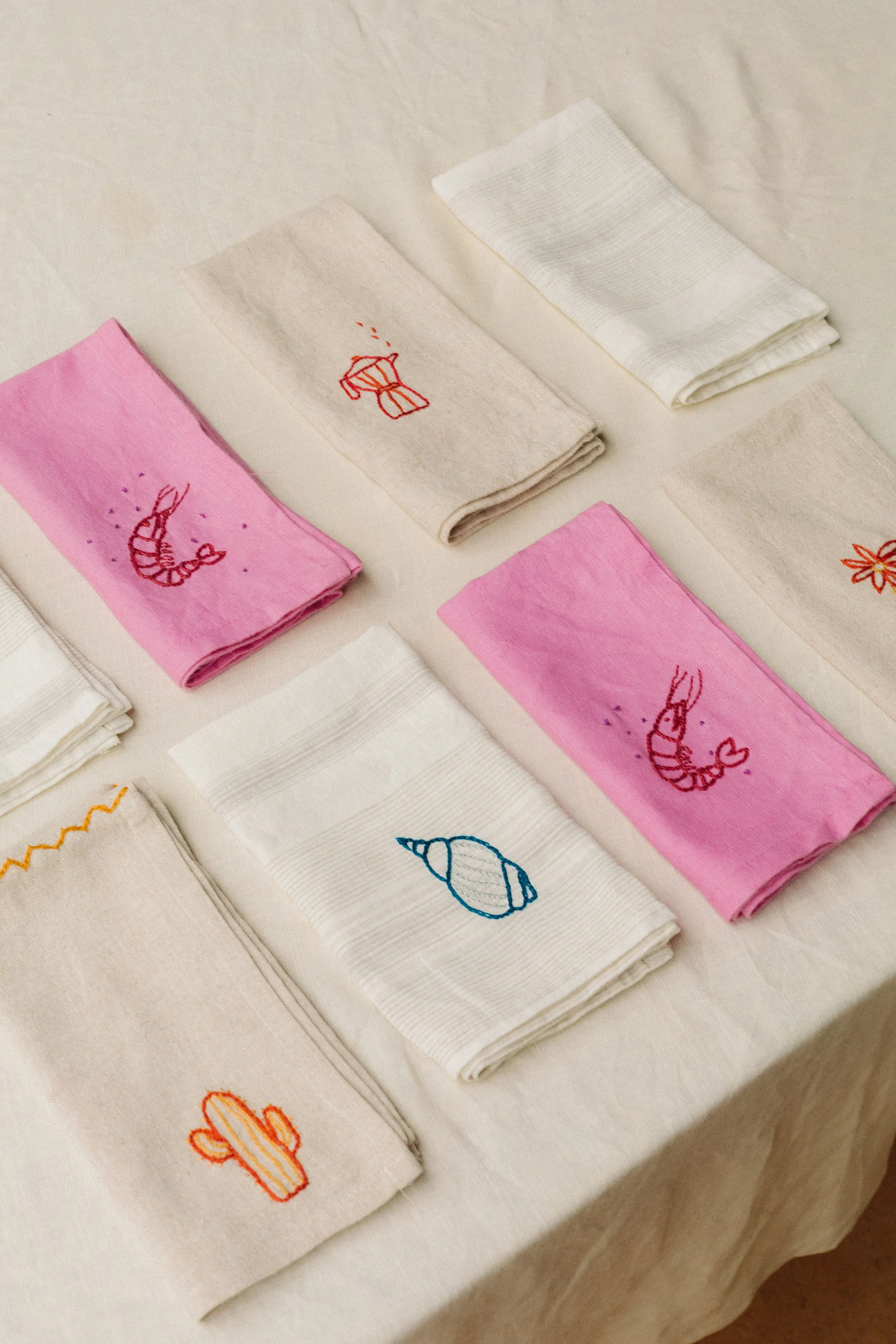 Stacked cloth napkins with embroidered summer motifs including a seashell, a potted plant, a flower, and an ice cream cone, in various colors such as red, blue, pink, and orange, displayed on a white table.