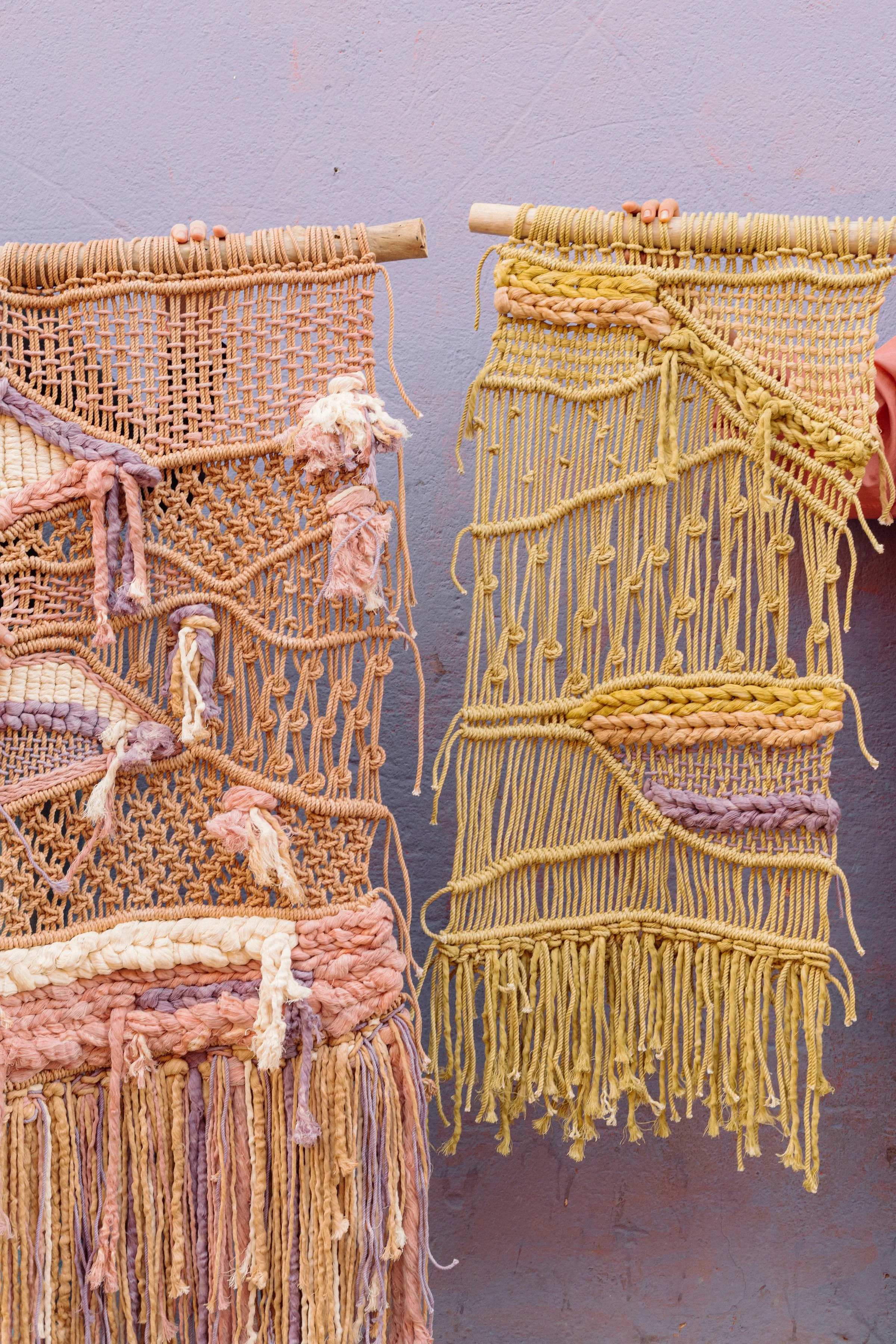 Colorful macrame wall hangings hanging on a metal rack outdoors, next to lush green trees and plants with white architectural features in the background.