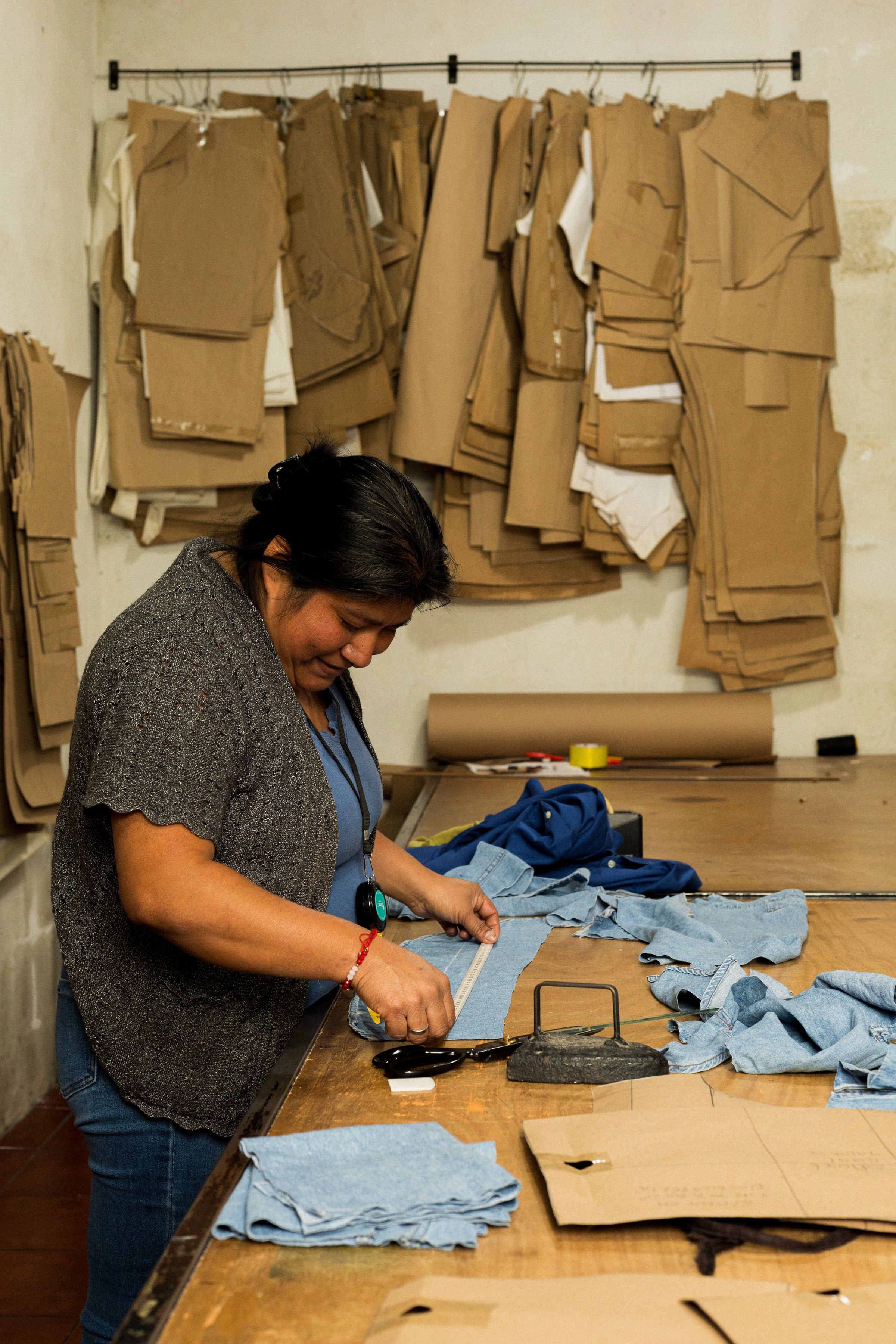 A woman working in a sewing or tailoring workshop, cutting fabric on a large wooden work table, with multiple stacks of fabric and pattern paper around her, and cardboard pattern templates on the wall behind her.