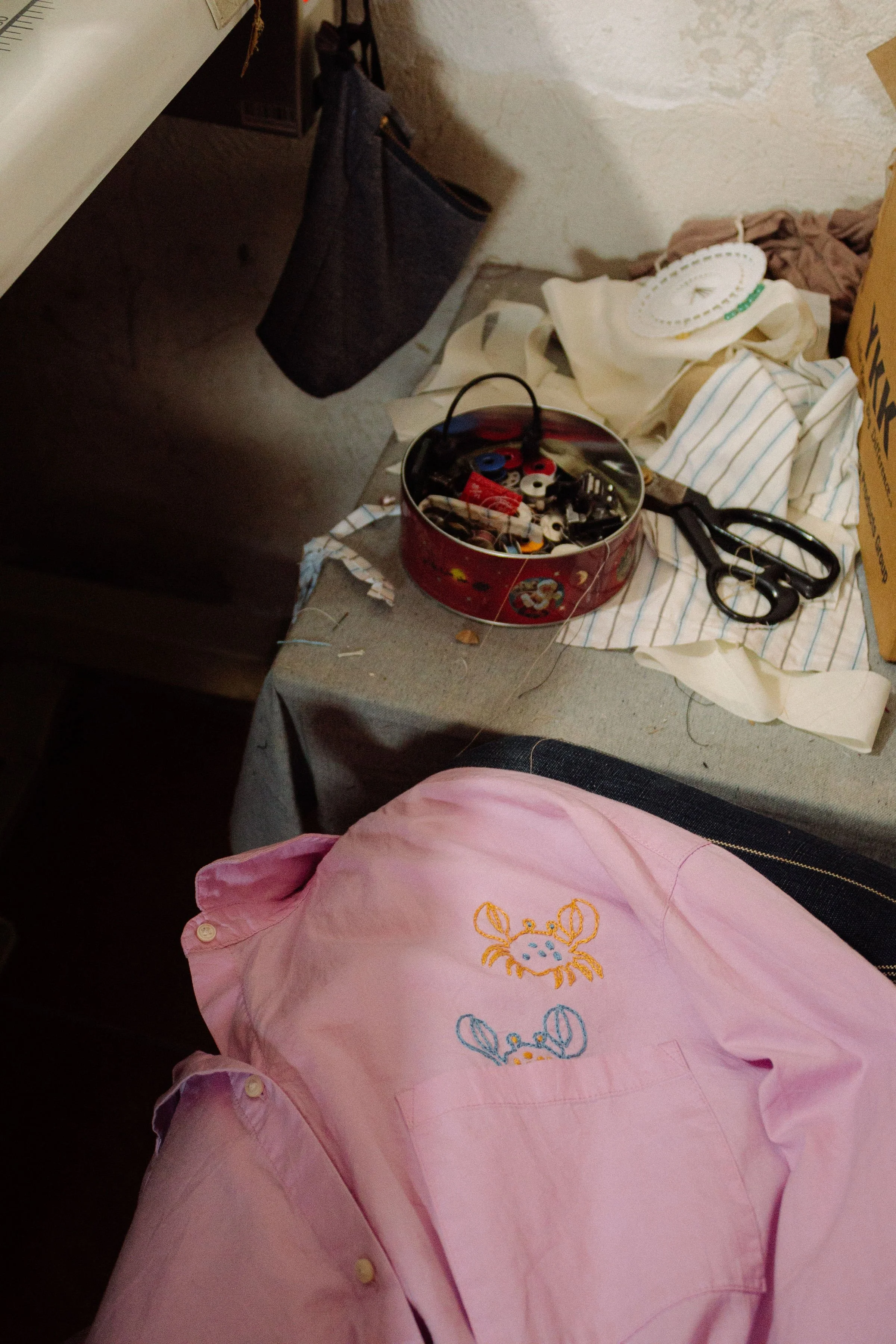 A cluttered table with a pair of scissors, a round box filled with various electronic parts, and some clothing and fabric items. The background includes a black bag, a cardboard box, and a textured wall.