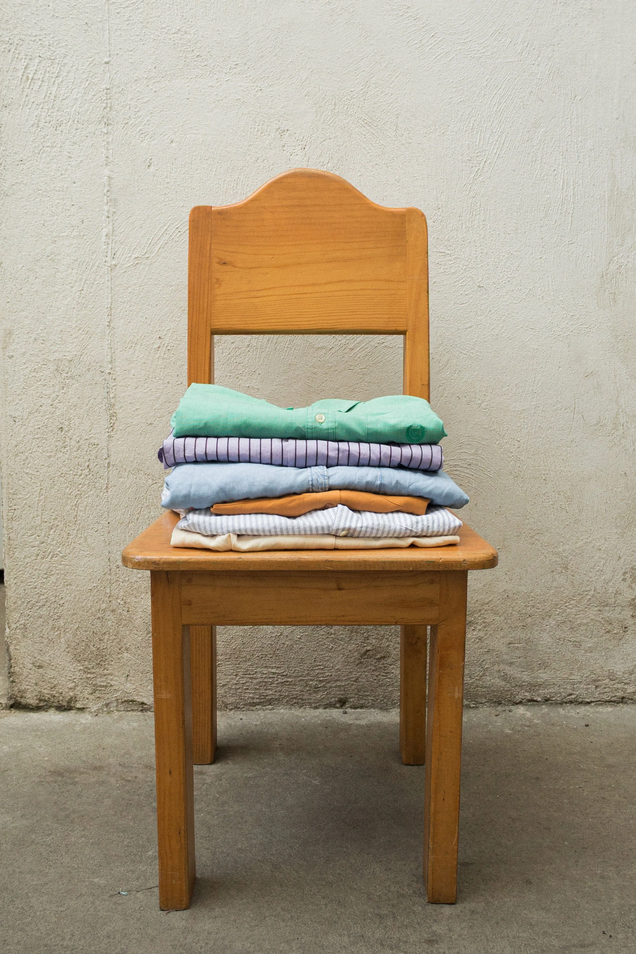 A wooden chair with a stack of folded clothes, including a green shirt, purple striped shirt, light blue jeans, beige pants, and a white shirt, placed on the seat, against a textured beige wall.