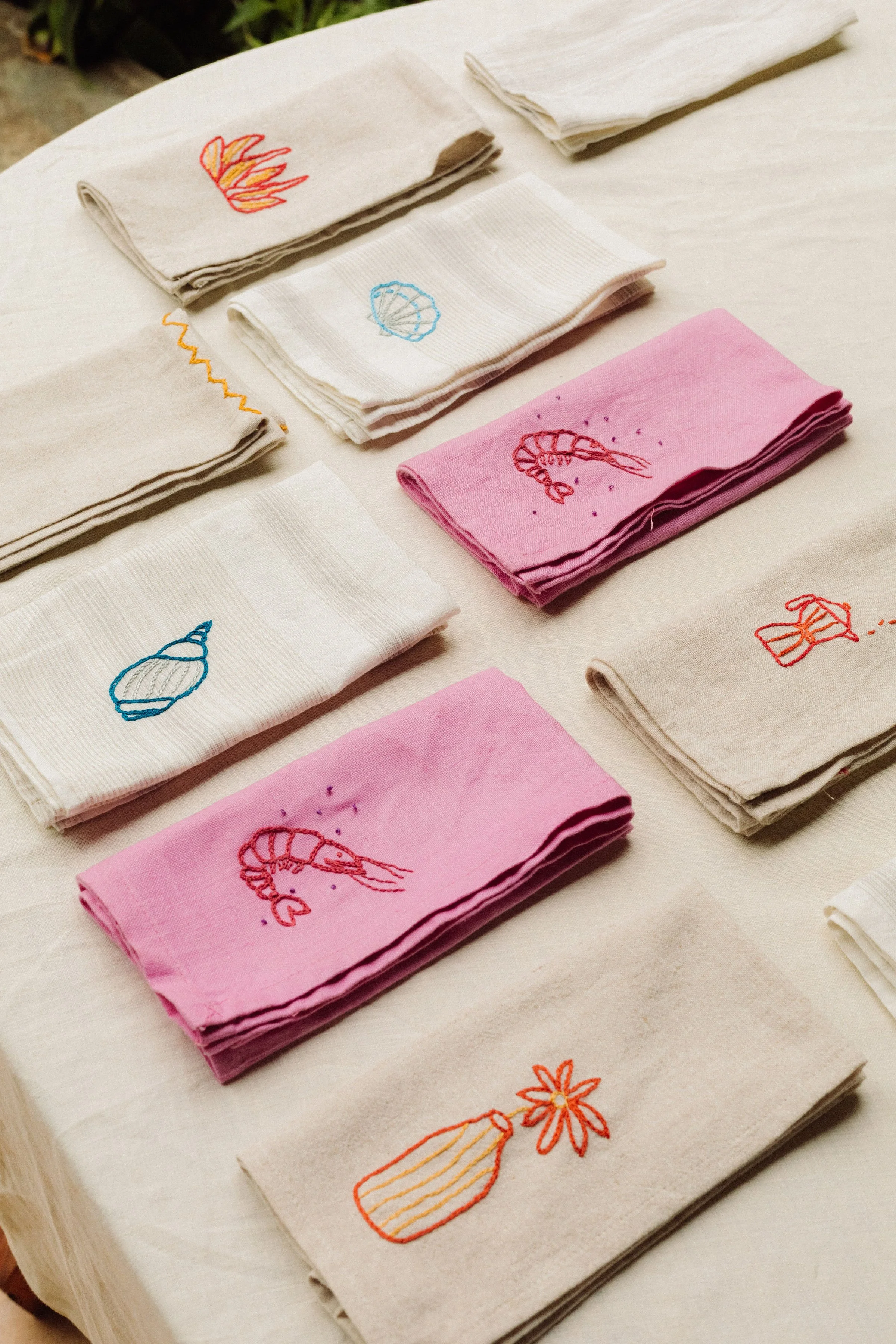 Cloth napkins with embroidered designs of seashells, coral, and flowers arranged on a white table.