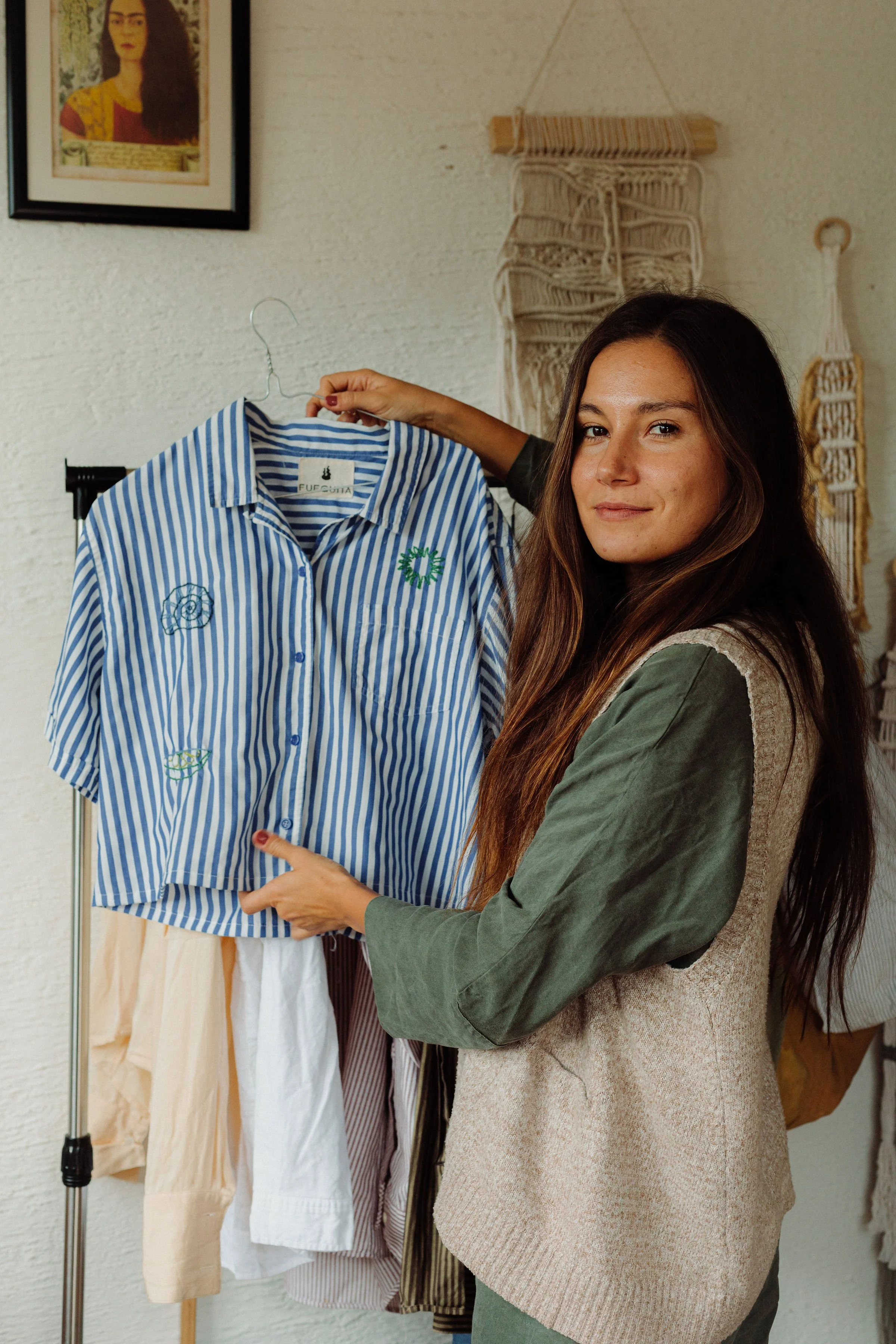 A woman with long brown hair is holding a blue and white striped shirt on a hanger and smiling at the camera in a room with beige and white clothing hanging in the background.