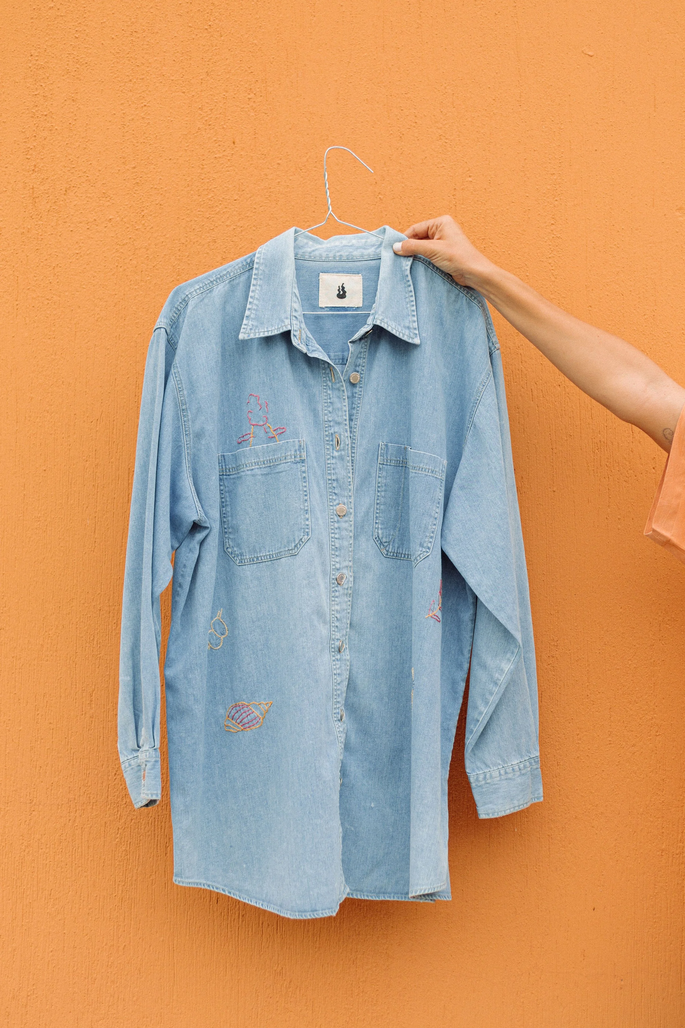 Light blue denim shirt hanging on a hanger against an orange wall, with embroidered designs of seashells and a girl fishing.