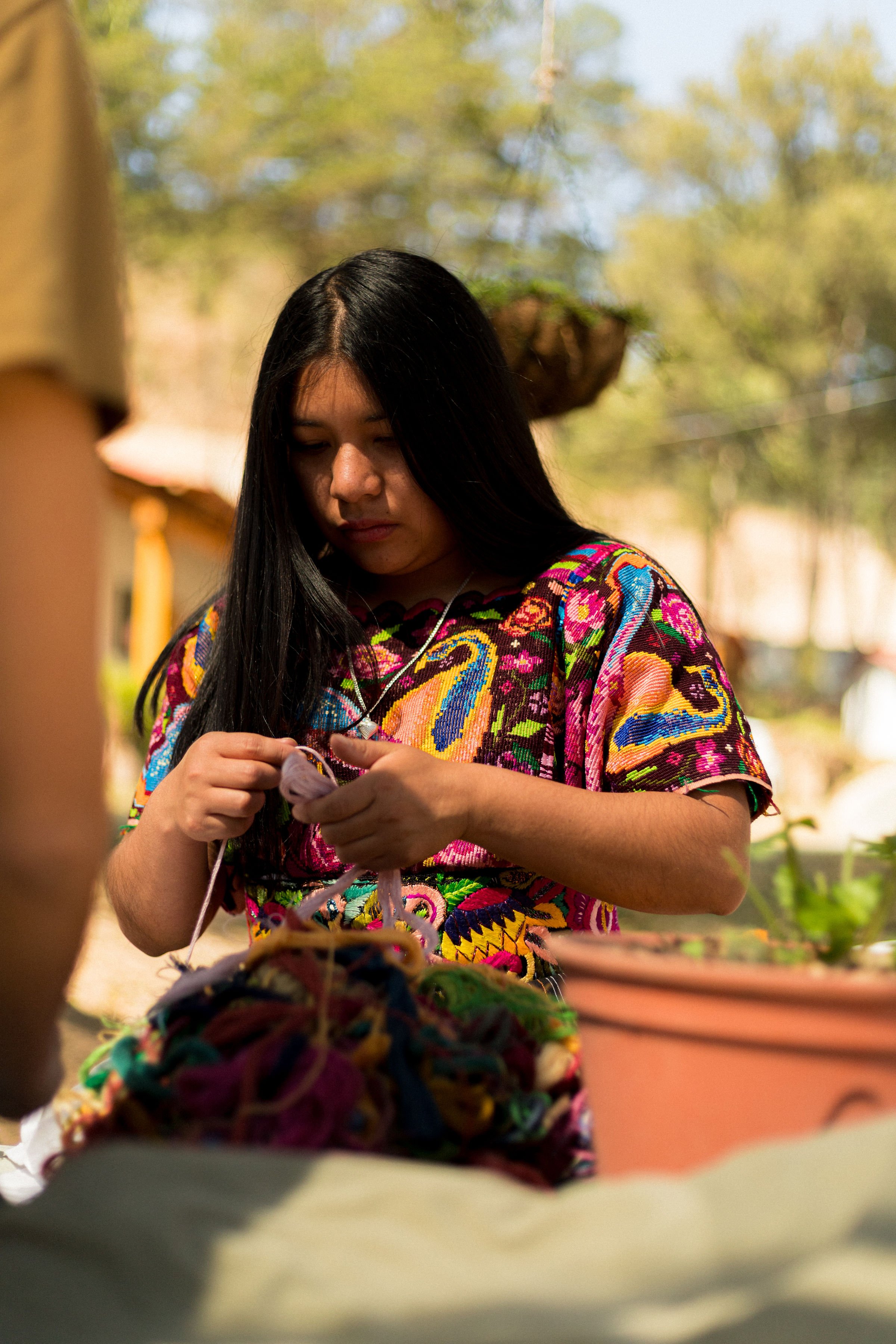 Young woman with long black hair wearing colorful embroidered blouse, knitting outdoors with natural background, possibly in a park or garden.