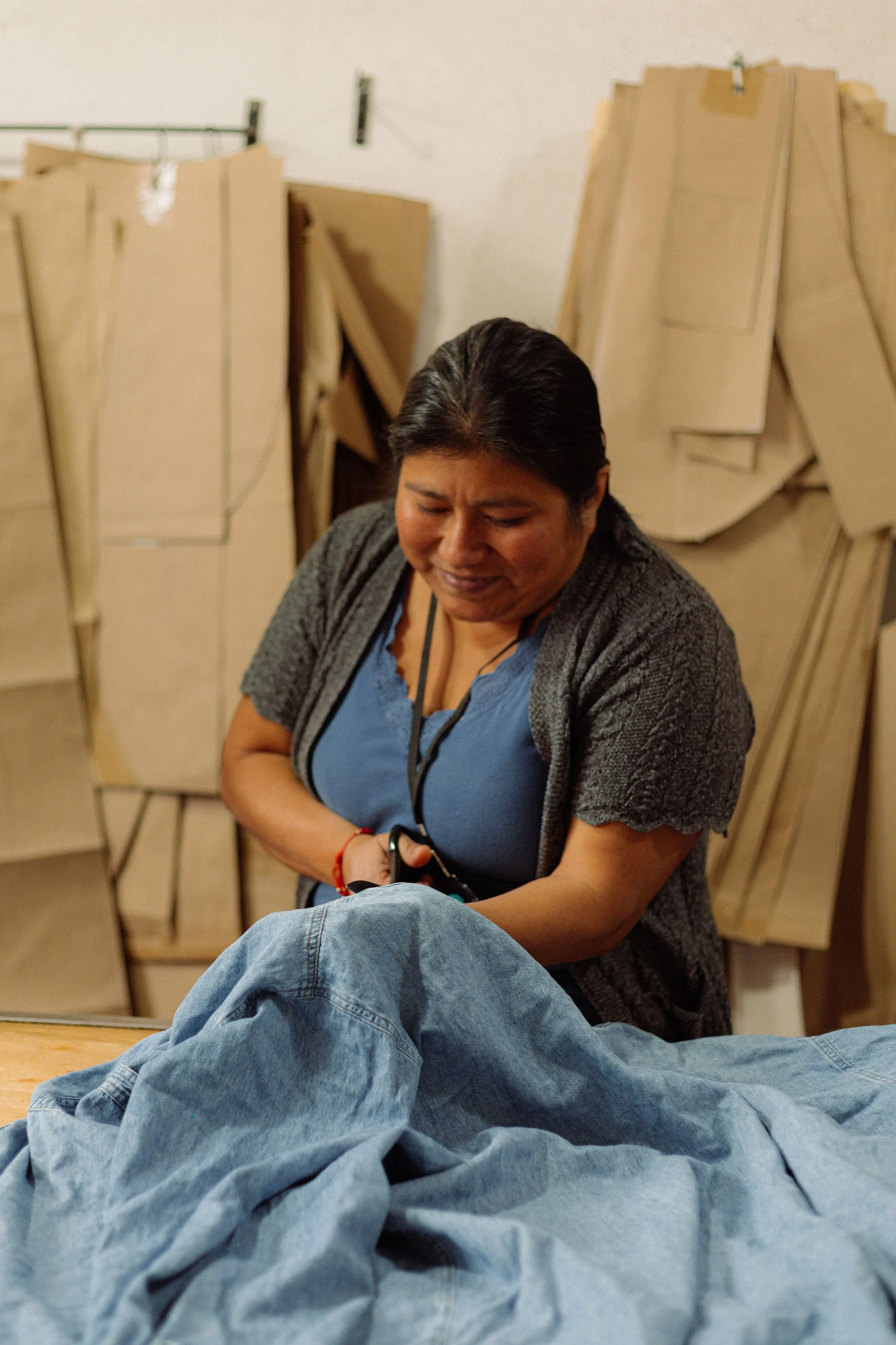 A woman with short black hair smiling, sitting in front of a line of wooden chairs, wearing a gray cardigan and a blue top, with a denim-clad person lying in front of her.