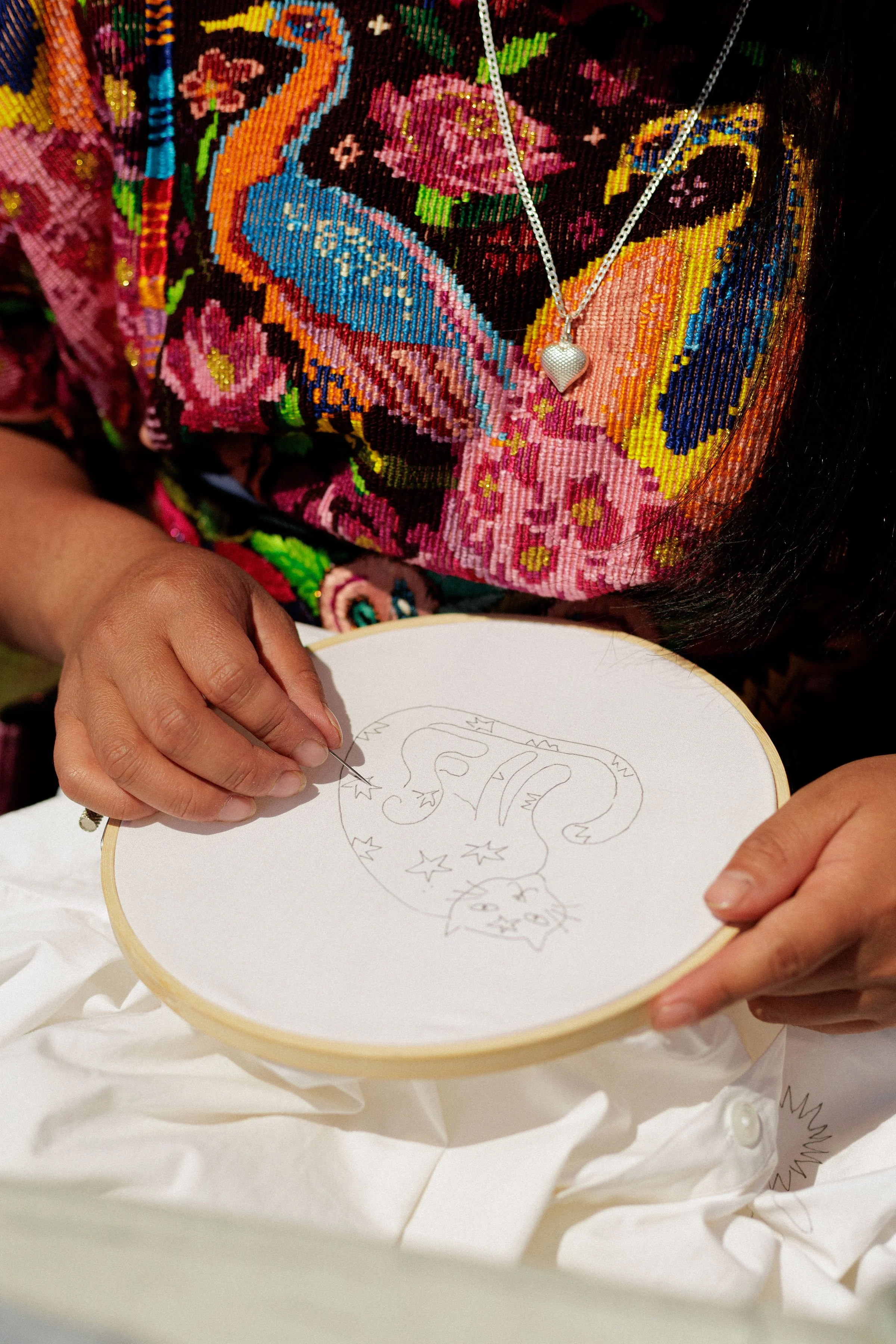 A person wearing a colorful, embroidered floral and bird-patterned garment is holding an embroidery hoop with a simple sketch of a lion drawn on fabric. The person is wearing a silver heart-shaped necklace.