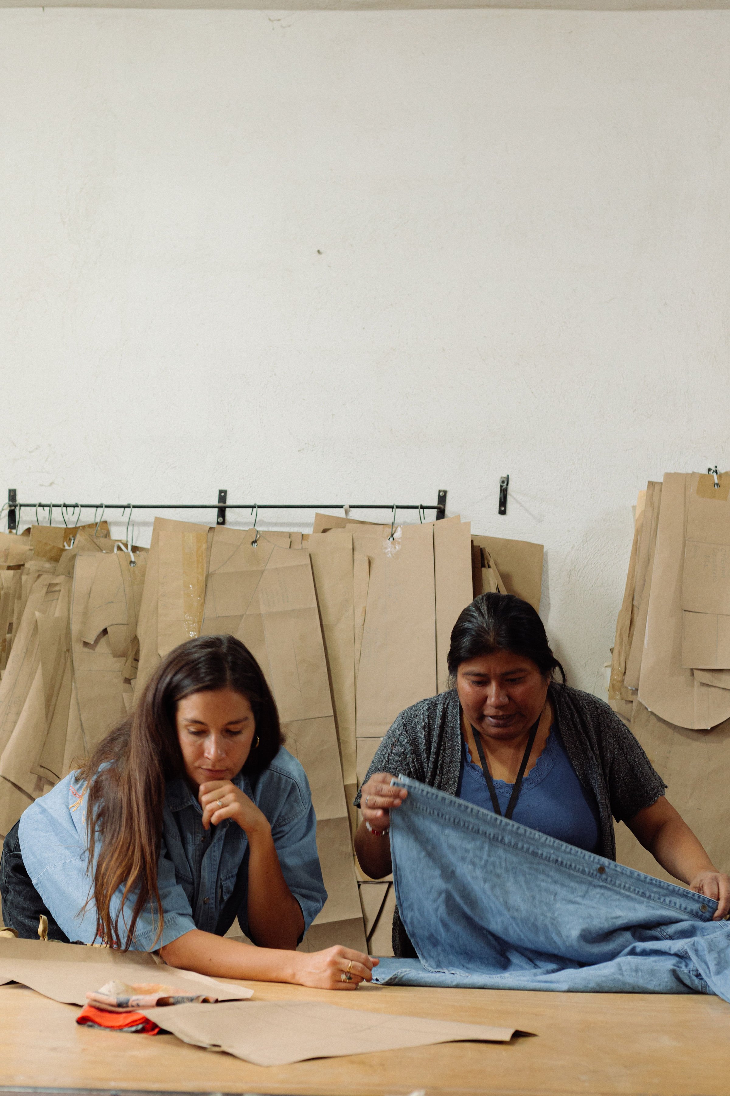 Two women sitting at a table, working on sewing projects with fabric and paper materials on the table, in front of a white wall and hanging paper bags in the background.