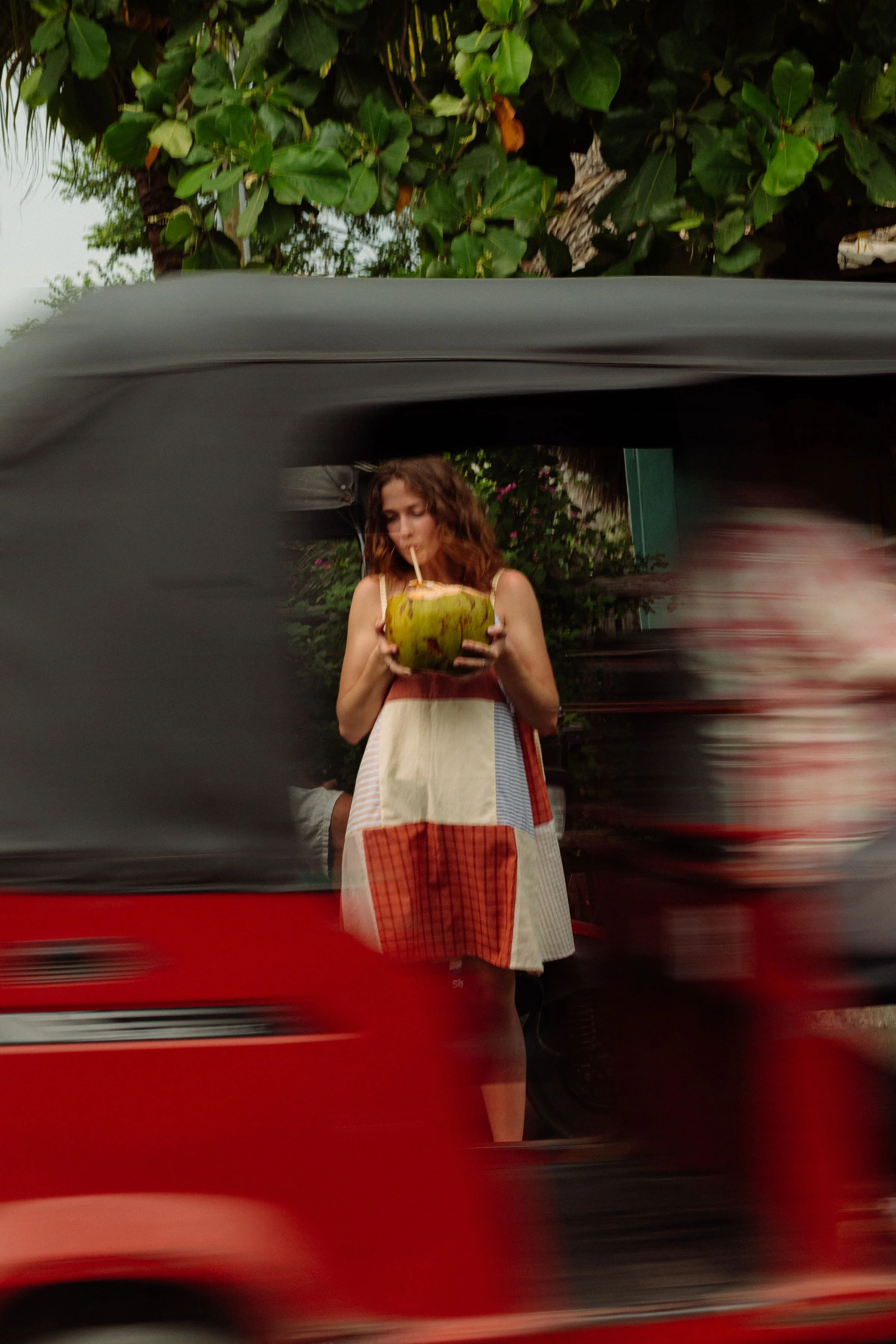 A woman with curly hair in a colorful dress holding a green coconut and drinking from a straw, standing outdoors surrounded by greenery.
