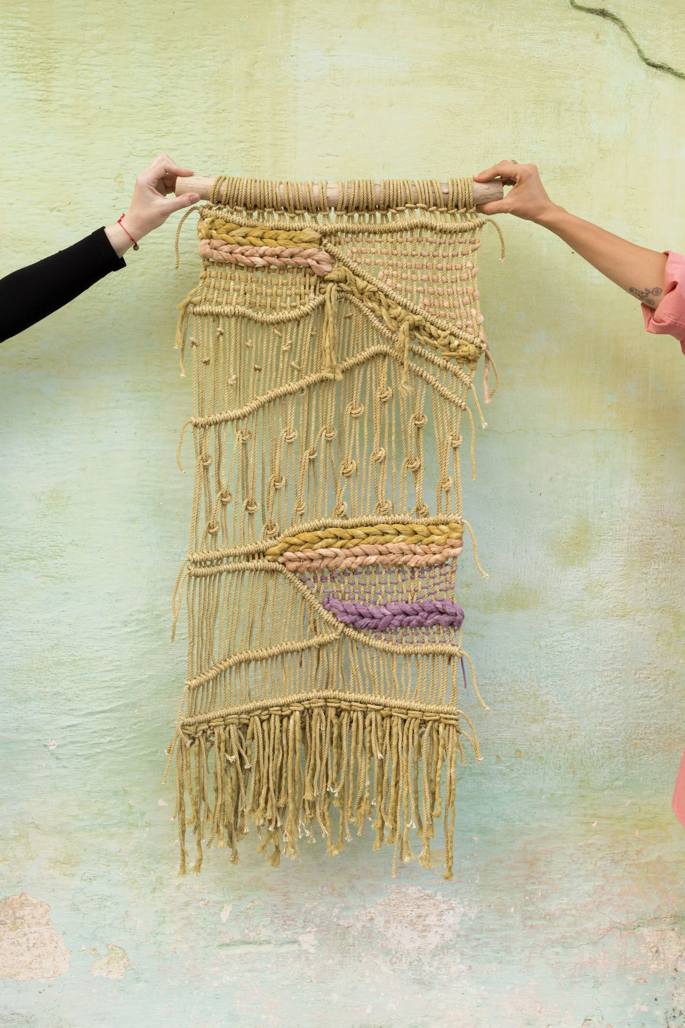 A textile wall hanging with various woven patterns and textures in shades of peach, beige, and cream, held up by a person's hands on a light gray wall.