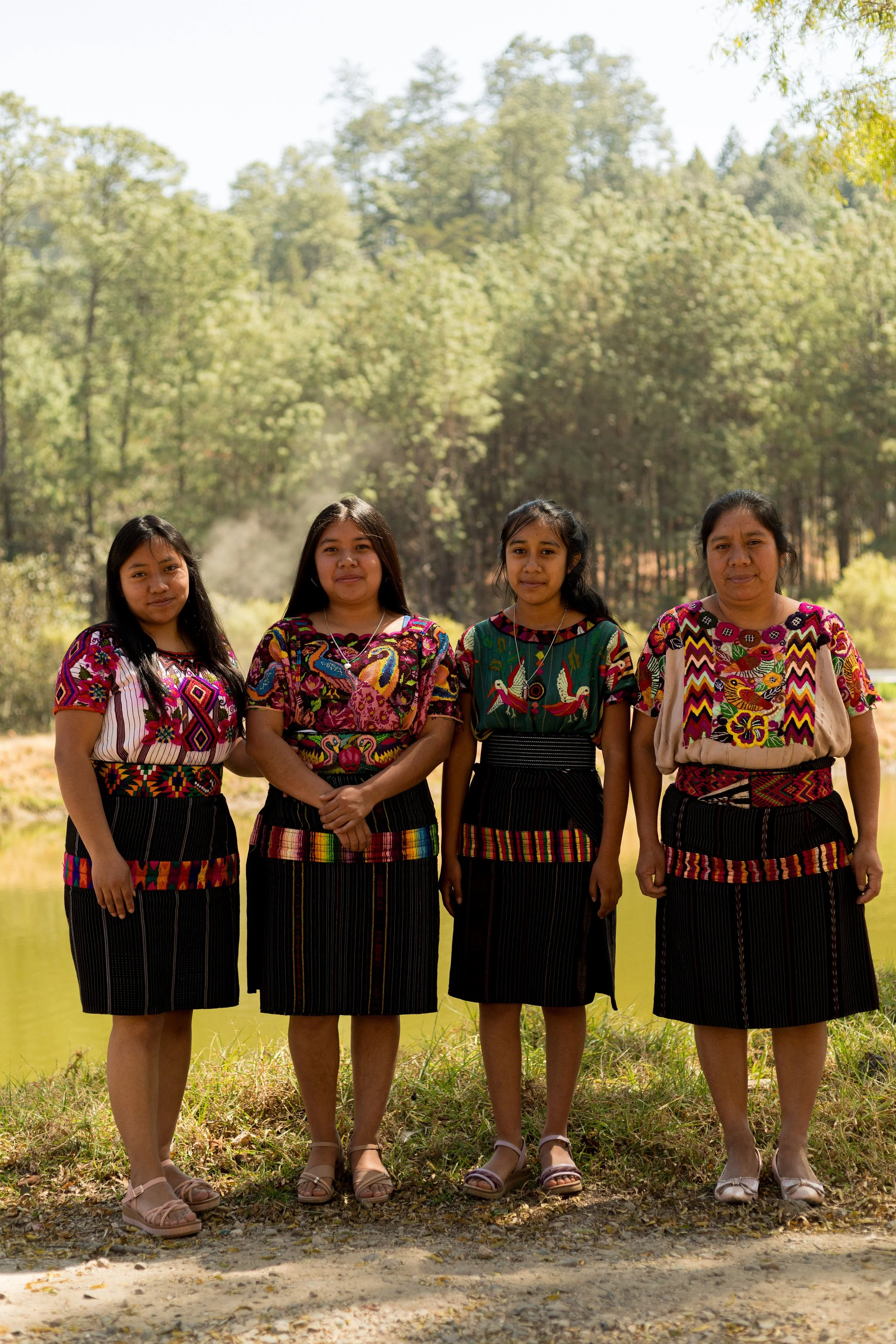 Four women standing in front of a lake surrounded by trees, wearing traditional colorful huipil blouses and black skirts, smiling at the camera.