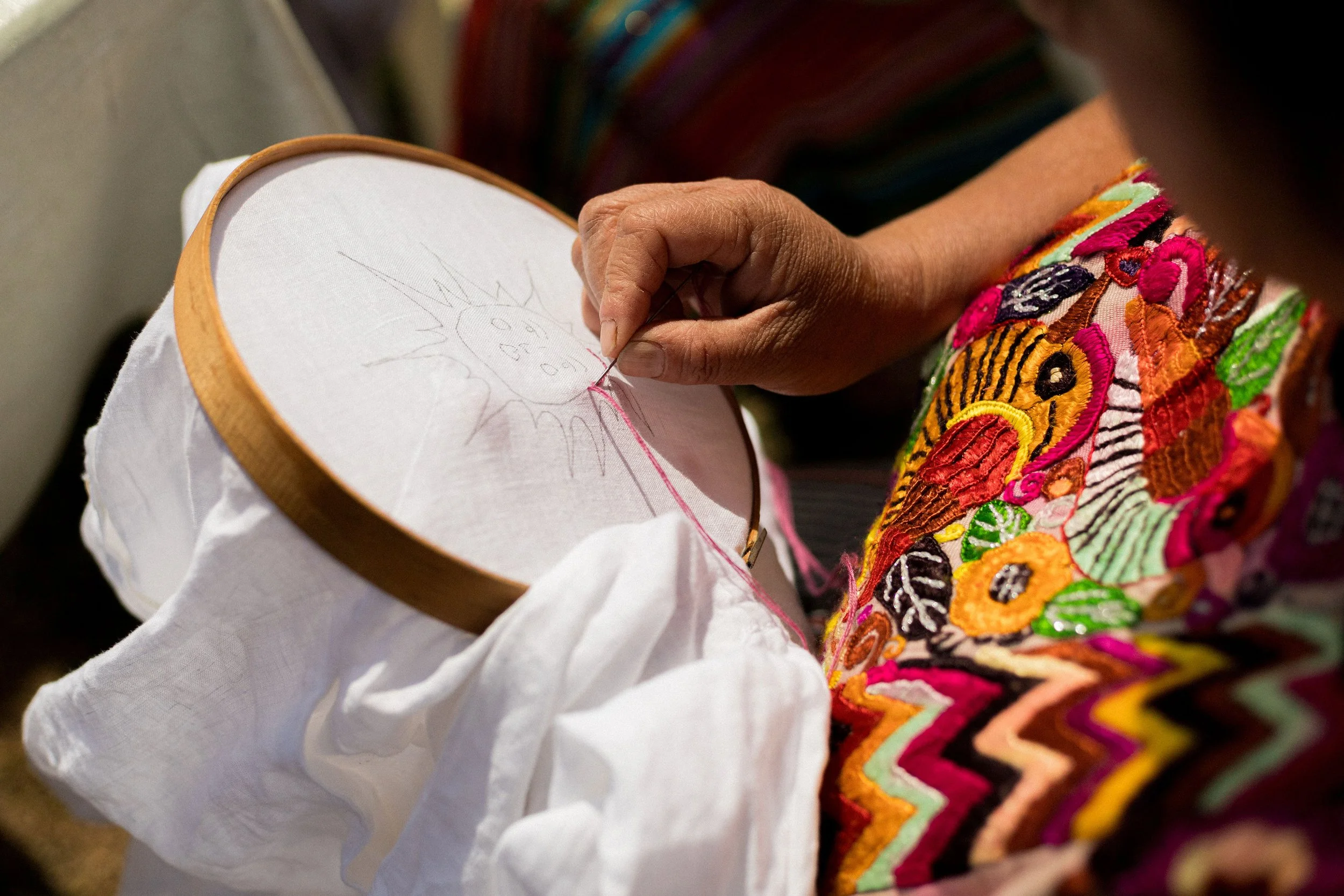 A person sewing colorful, intricate embroidery onto fabric, with a small embroidered sun sketch on paper nearby.