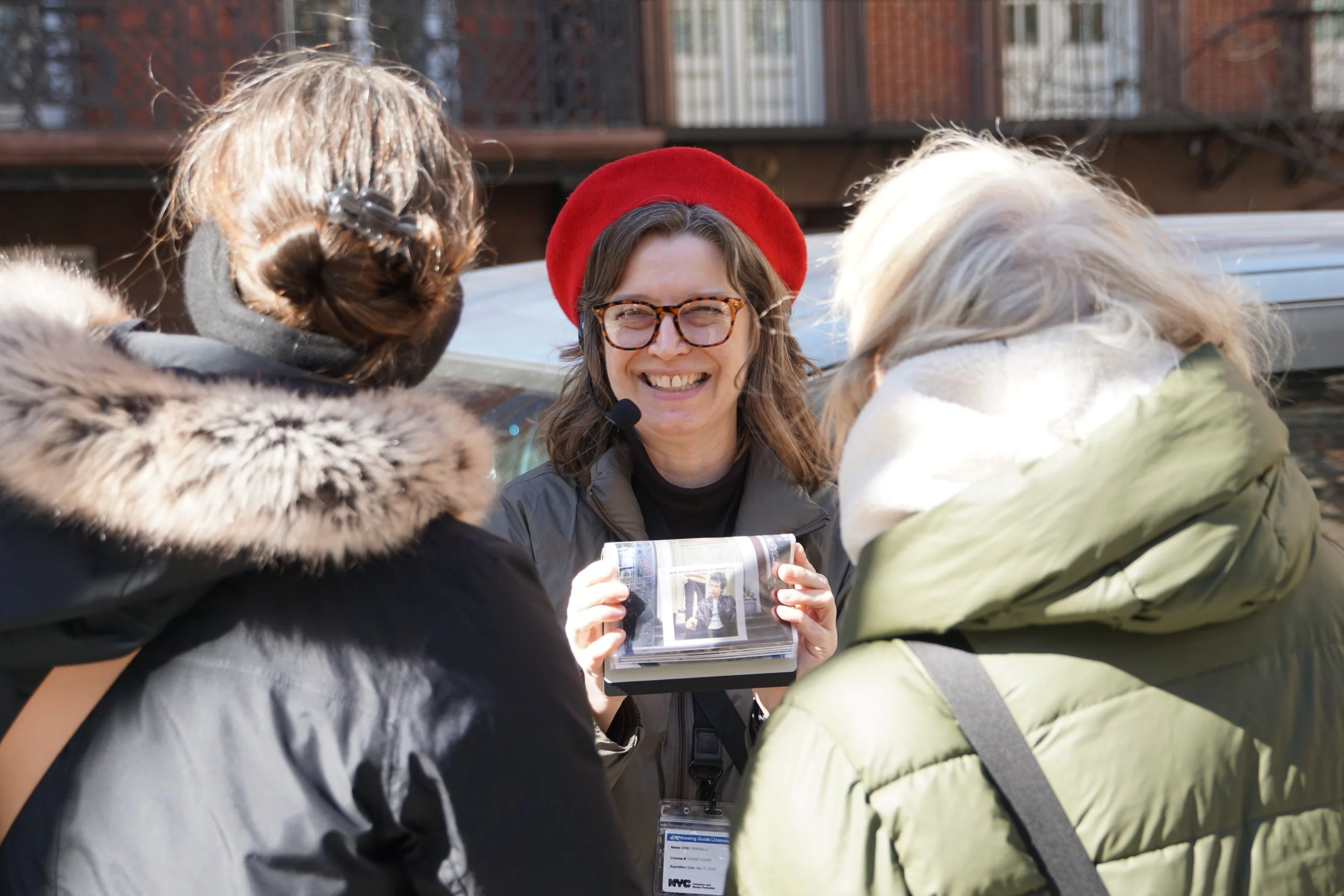 A person wearing glasses and a red beret, using a microphone headset, is smiling while holding a photo album. Two people with winter jackets, viewed from behind, are looking at them. The background shows a building and a car.