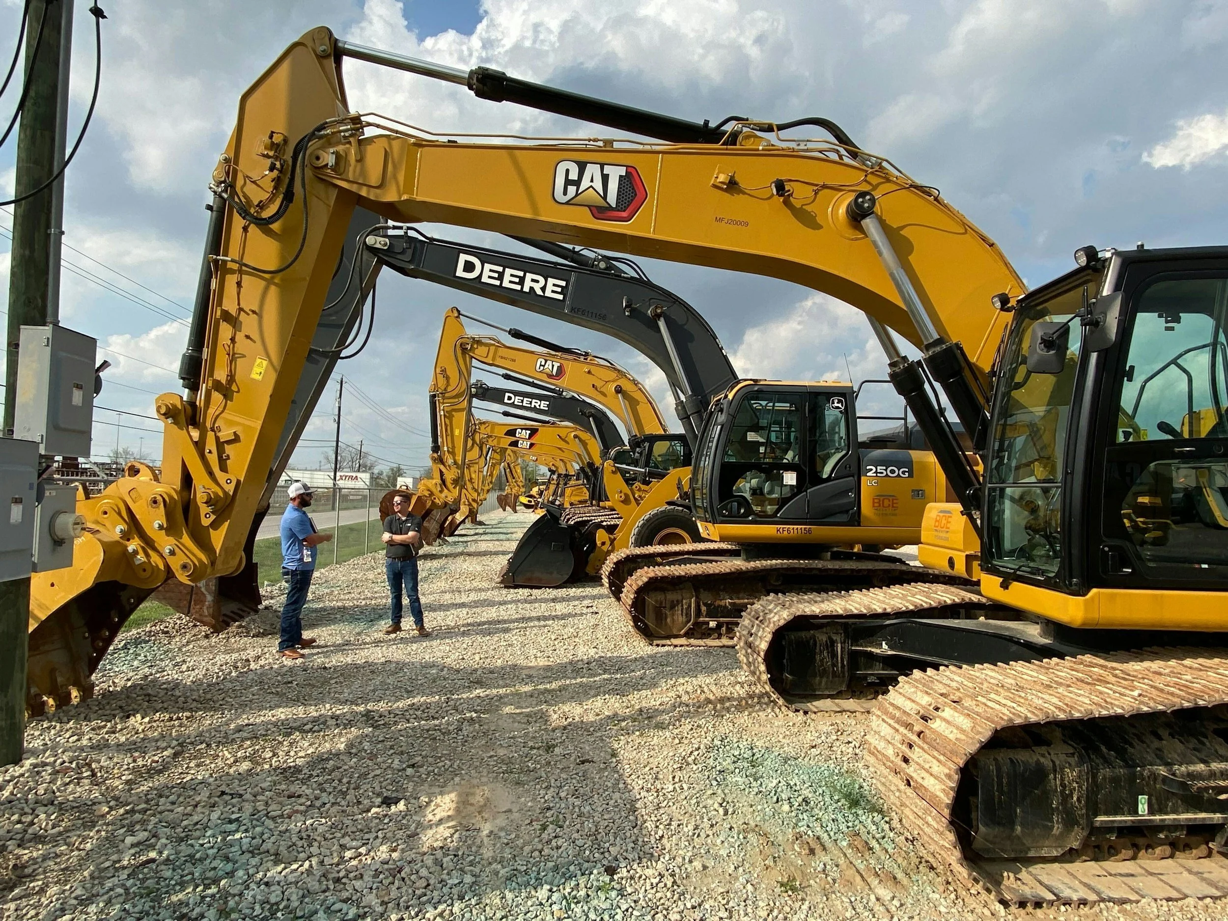 Multiple yellow construction excavators with brand logos, parked on a gravel lot, with a few people inspecting them and a cloudy sky above.