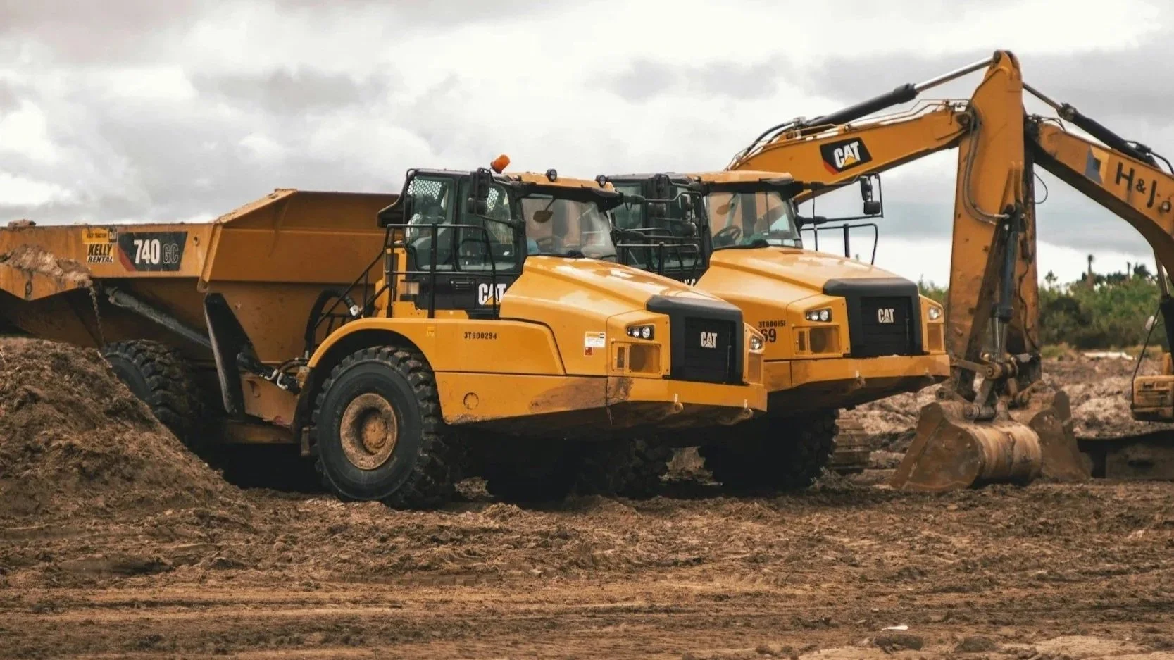 Two yellow Caterpillar construction dump trucks and an excavator working on a construction site with dirt and overcast sky.
