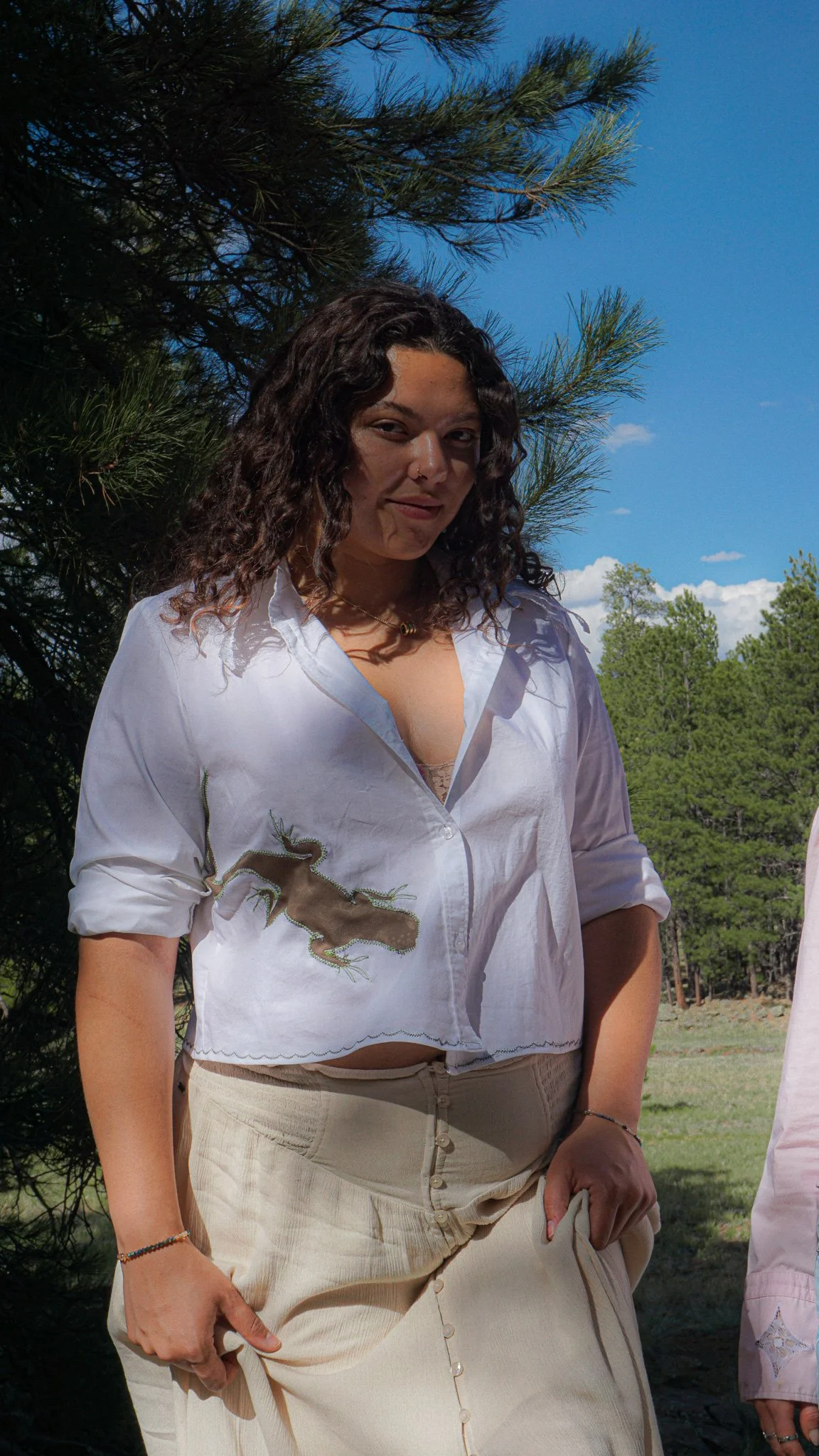A woman with shoulder-length hair wearing a bright pink T-shirt with graphic prints, dark shorts, white sneakers, and white socks, standing on a dirt or volcanic sand surface in front of a backdrop of green pine trees and mountains.