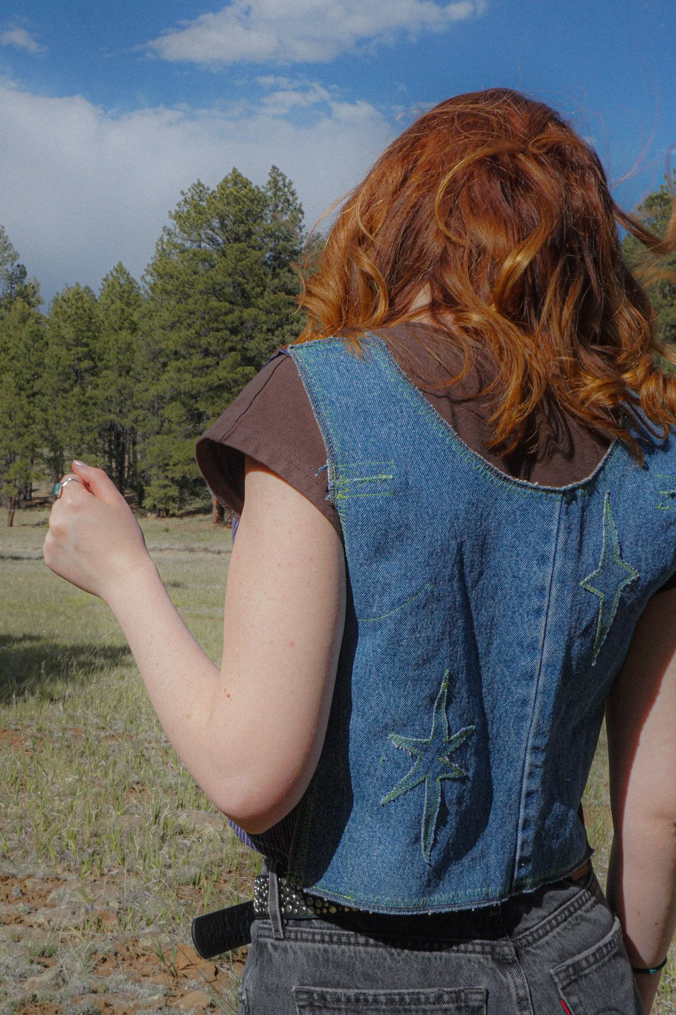Side profile of a young woman with wavy reddish-brown hair in an outdoor setting with a bright blue sky and green pine trees.