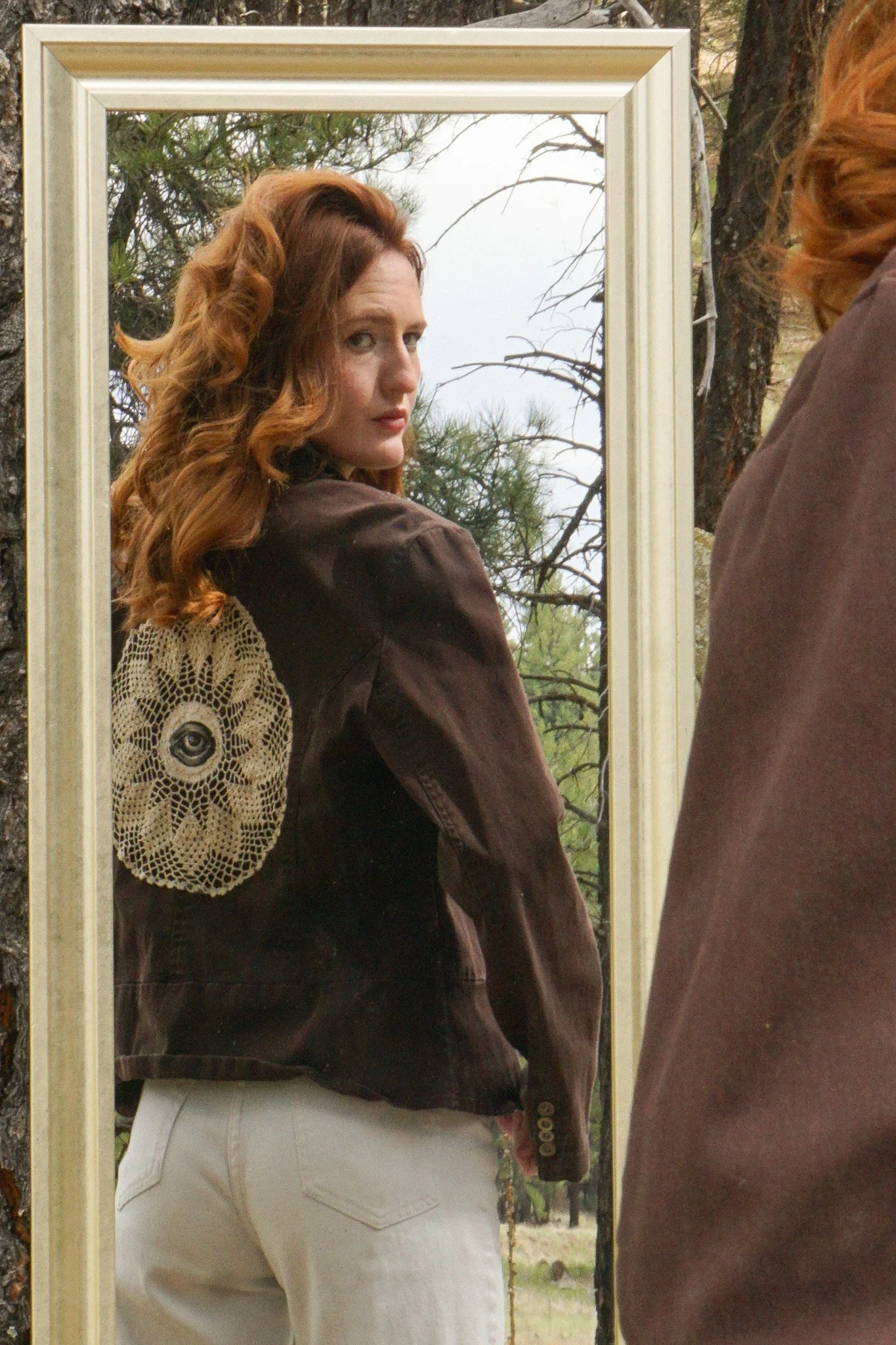 A person wearing a white t-shirt with black designed skeleton bones, holding a dried plant, sitting in a snowy field with tall dry grass and snow-capped mountains in the background under a clear blue sky.