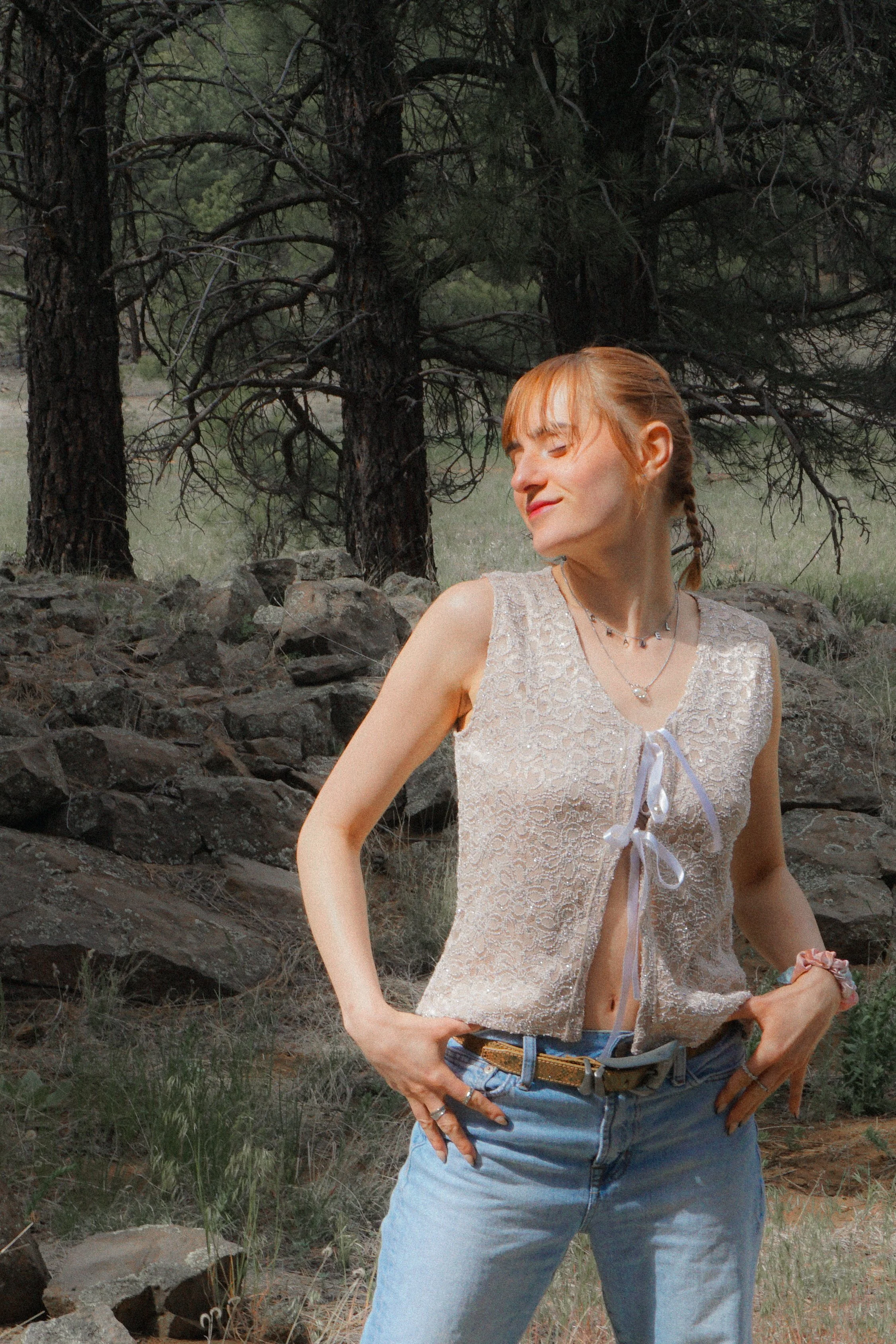 A young woman with light skin, wearing a black shirt with a large, colorful embroidered skeleton face and heart design, light blue jeans, and jewelry including large hoop earrings, a septum piercing, and layered necklaces. She is standing outdoors against a rustic wooden fence with her eyes closed and her face tilted slightly upward, with sunlight illuminating her face.