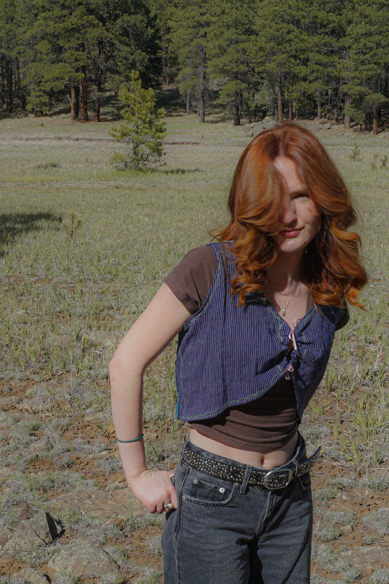 A woman with blond hair wearing a blue sweater with a graphic design, kneeling on the ground in a forested area with pine trees and dry grass, looking down with her eyes closed.
