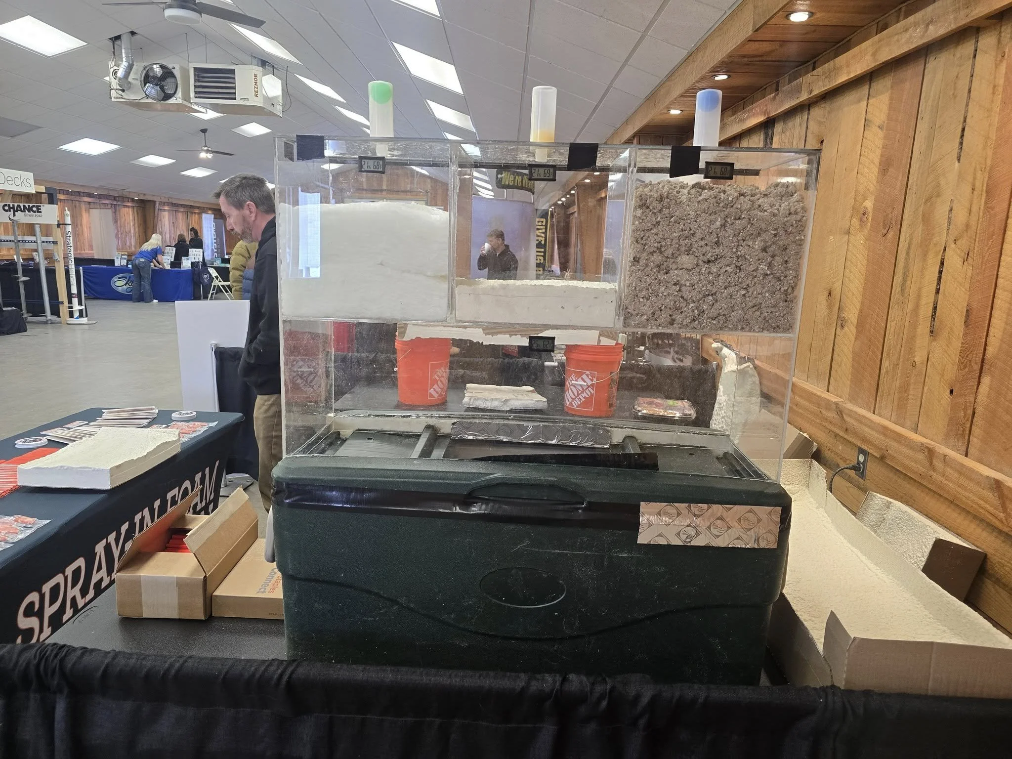 A display of different types of dry bulk ingredients in a plastic case at an indoor event, with people and booths visible in the background.
