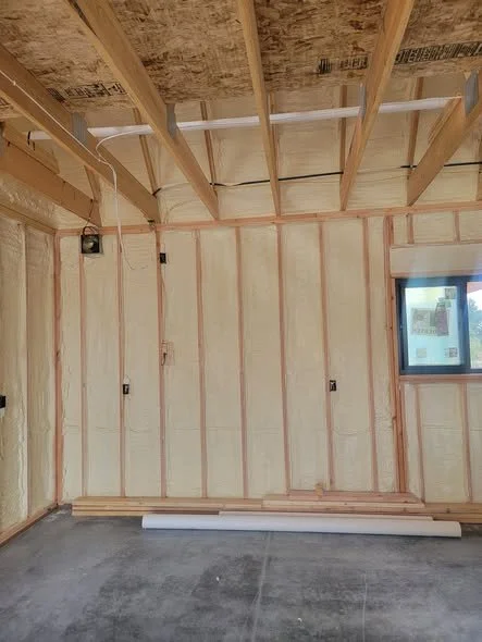 Interior of a room under construction with exposed wall studs, ceiling beams, and electrical conduit. There is a small window on the right side and construction materials on the floor.