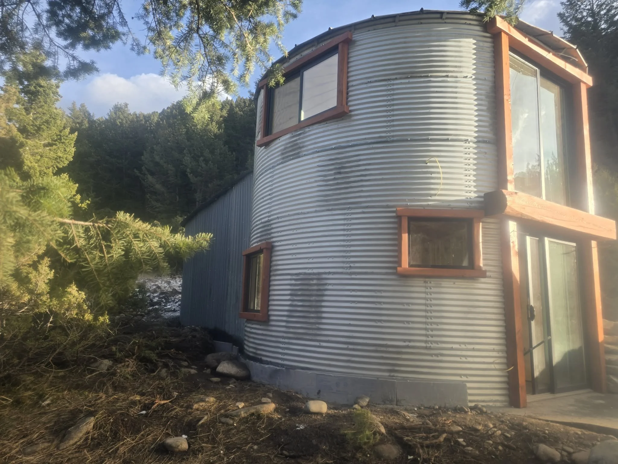 A rounded house constructed with corrugated metal siding, featuring wooden window and door frames, surrounded by natural landscape and trees with a mountain in the background.