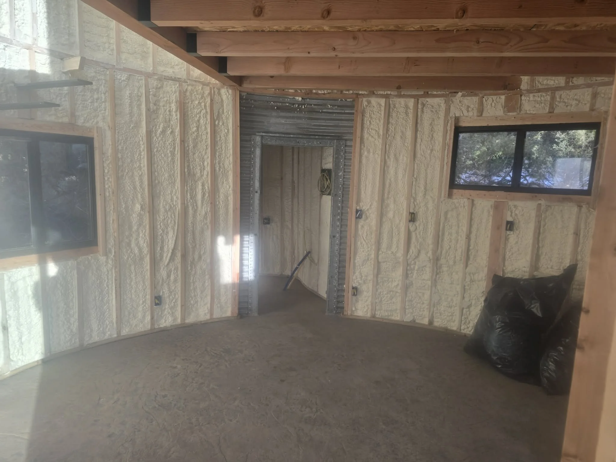 Interior of a room under construction with insulation on the walls, windows, and a doorway leading to another space. The floor is covered with dirt or construction material, and there are black garbage bags in the corner.