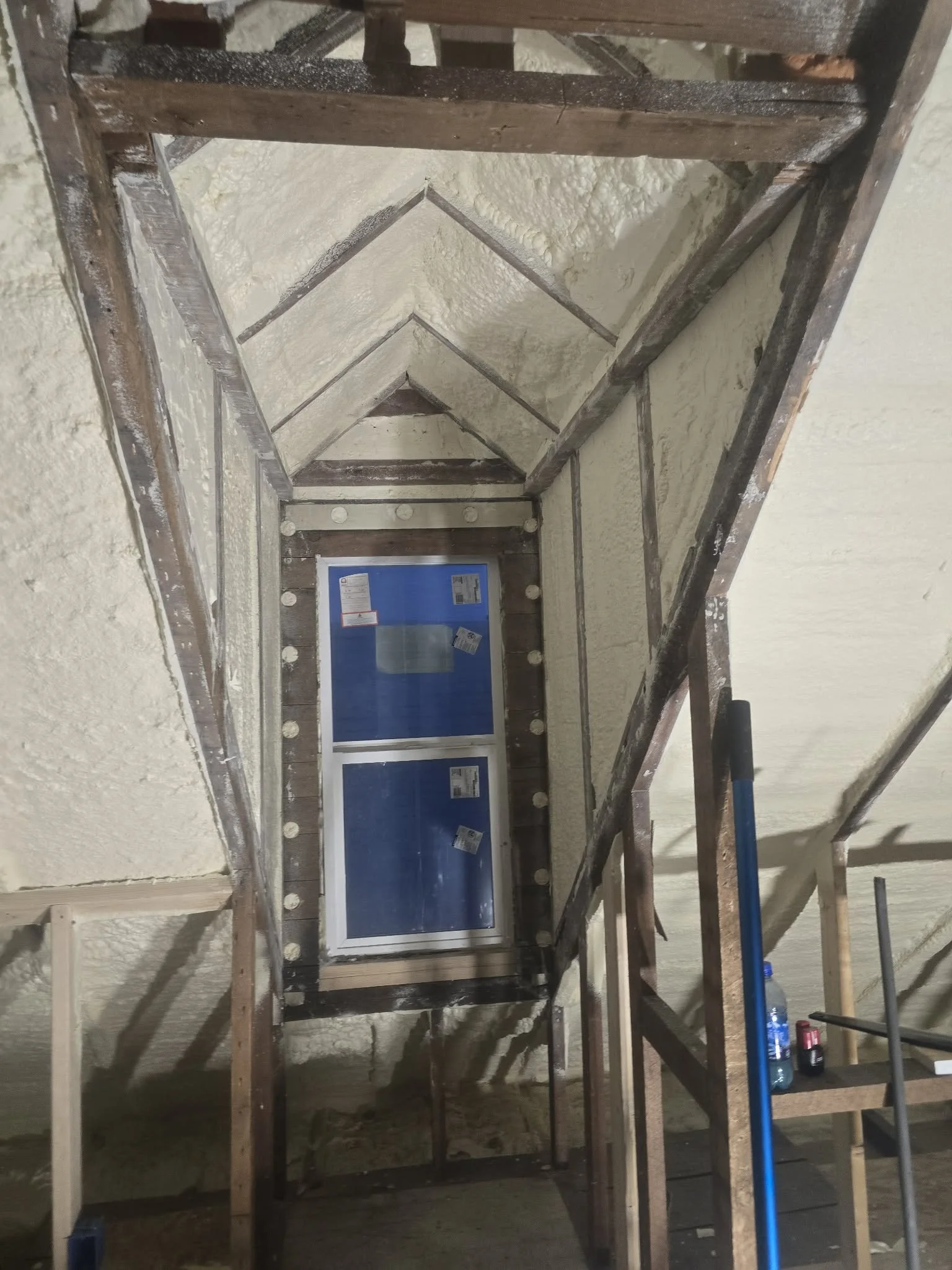 Interior view of attic or loft space under construction with a small window, wooden framing, and spray foam insulation on the roof.