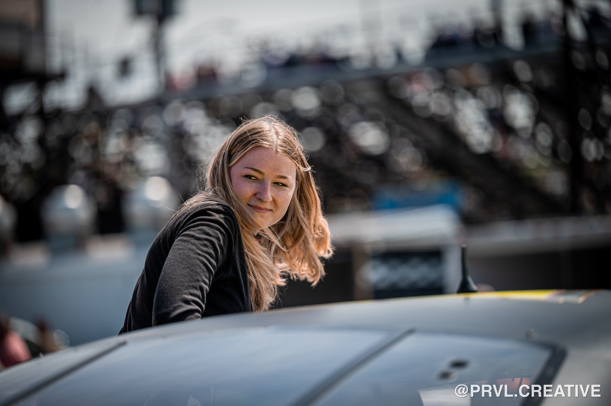 Young woman with blonde hair smiling partially hidden behind a silver car, with a background of boats and marina.