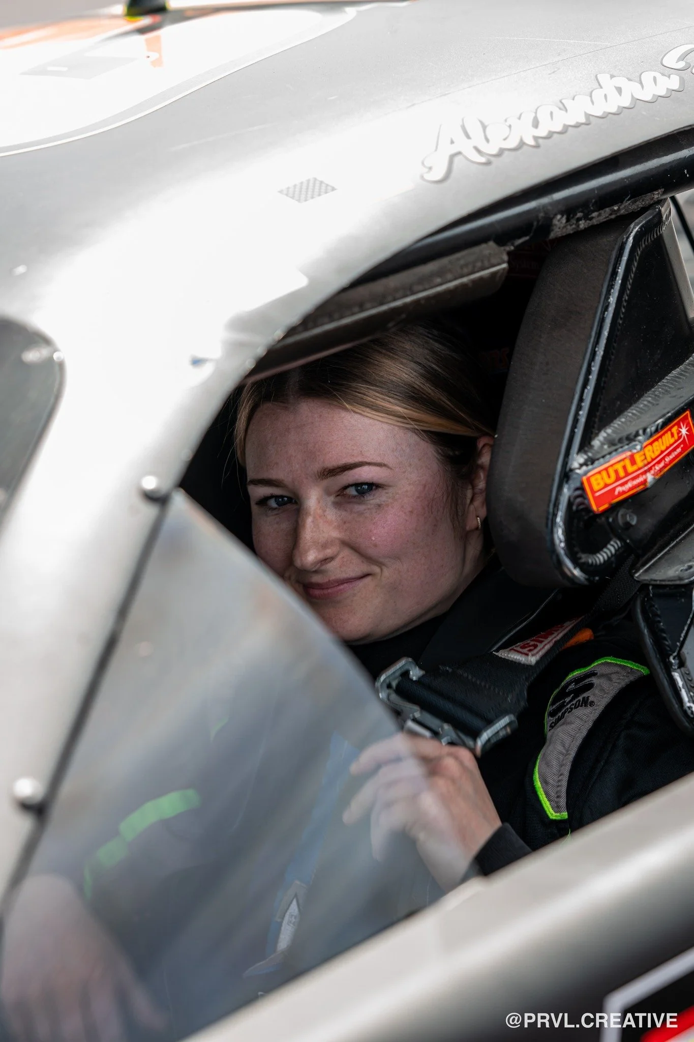 Close-up of a female race car driver sitting in the cockpit of her race car, looking out from the window with a slight smile.