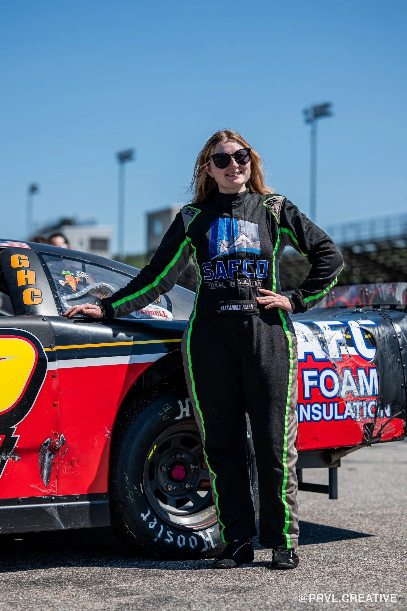A woman in a racing suit with sunglasses standing next to a race car on a track under a clear blue sky.
