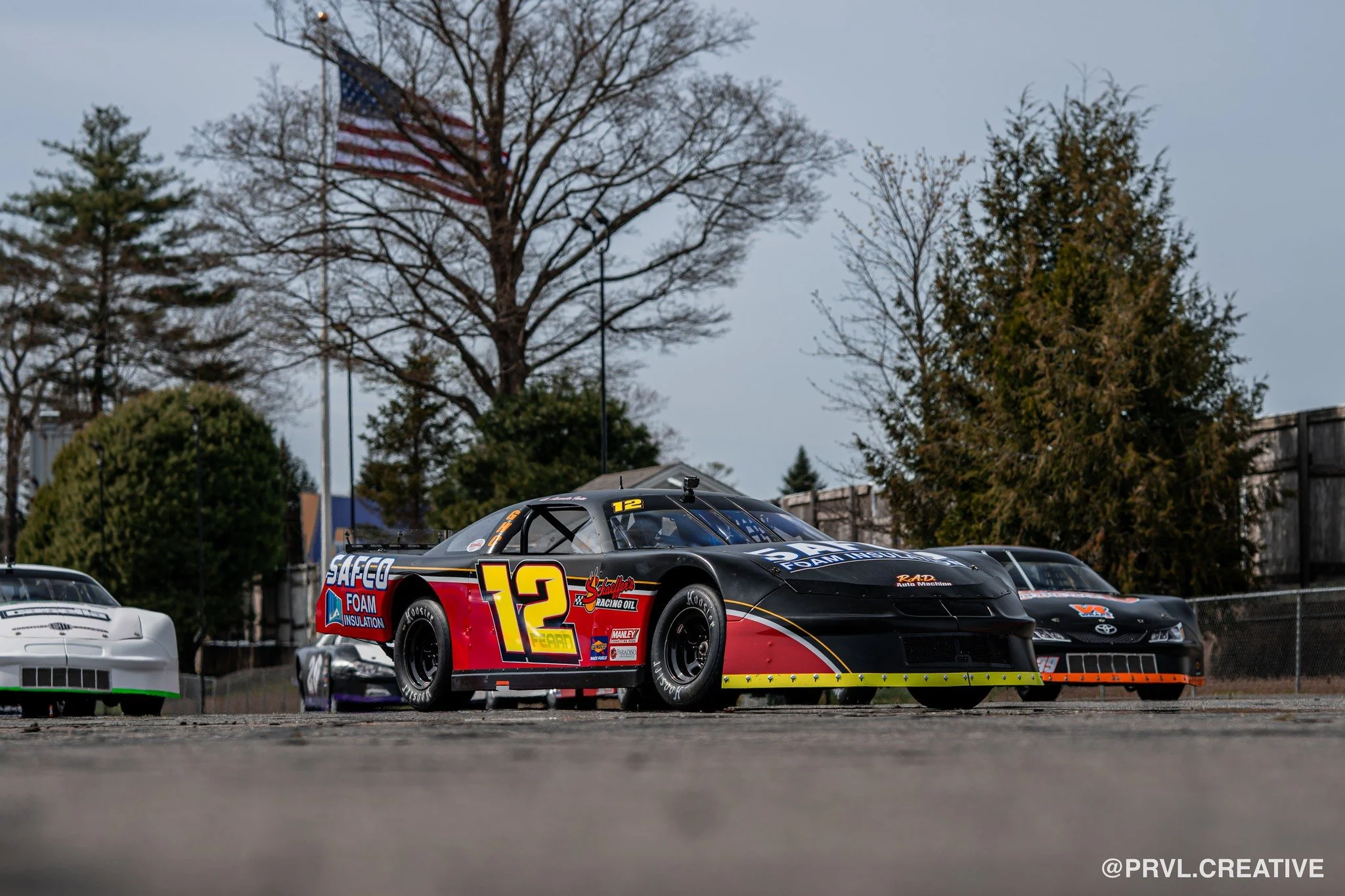 Race cars parked on a track with trees and an American flag in the background.