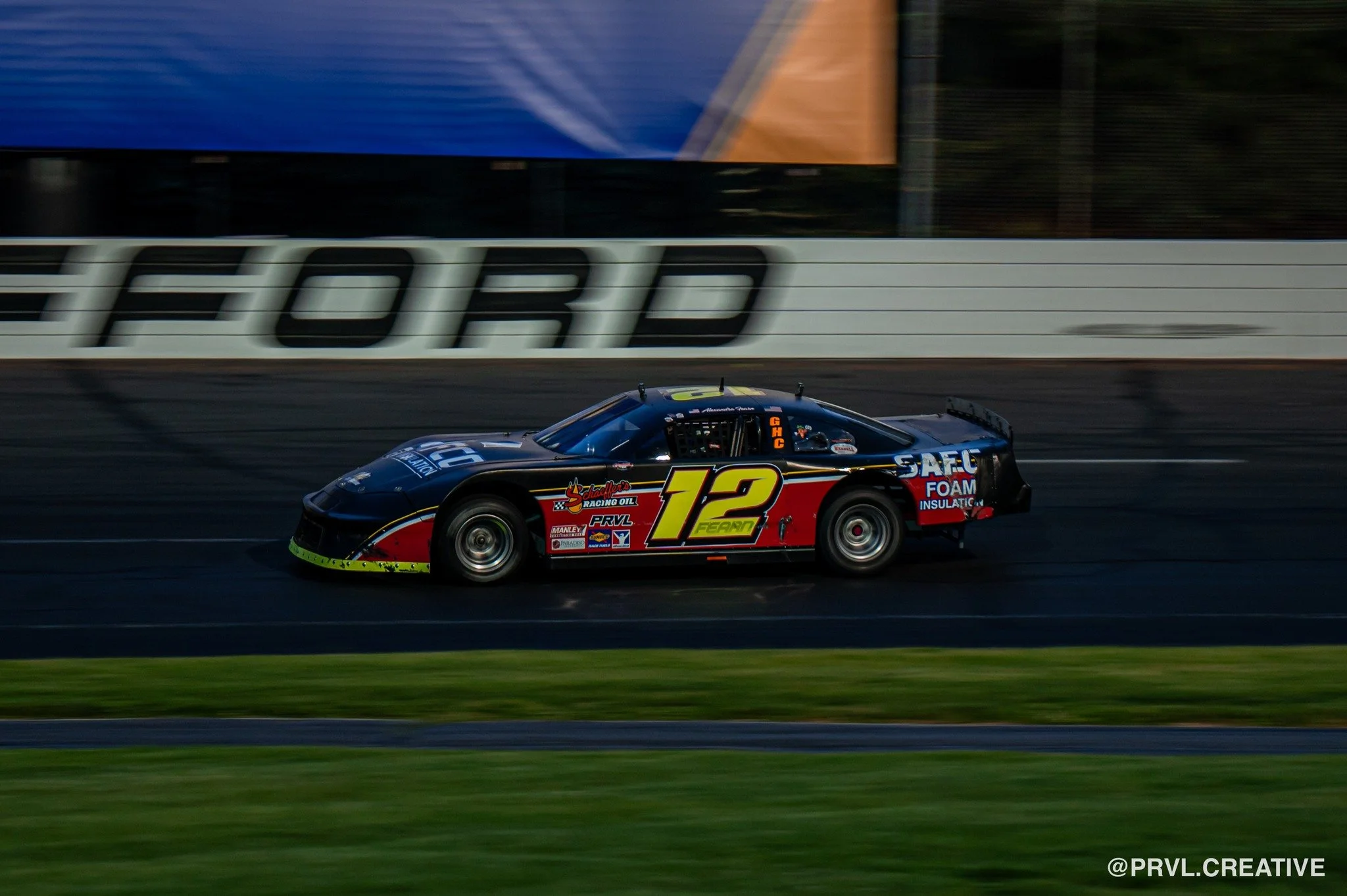 A black race car with the number 12 on its side racing on a track, with a blurred background showing the word 'HORD'.