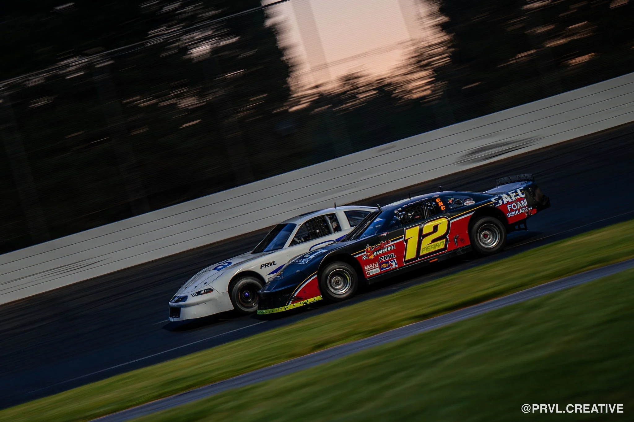 Two race cars on a track, one black with yellow and red number 12, and the other white, racing side by side at sunset.