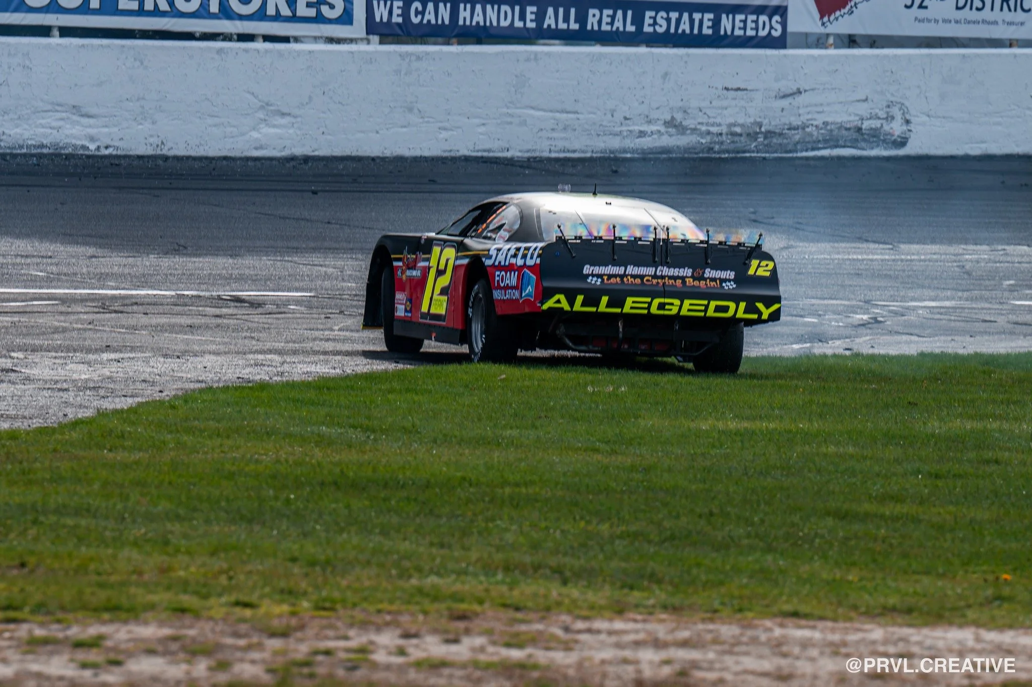 Race car on a track with grass in the foreground and ads on a white wall in the background.