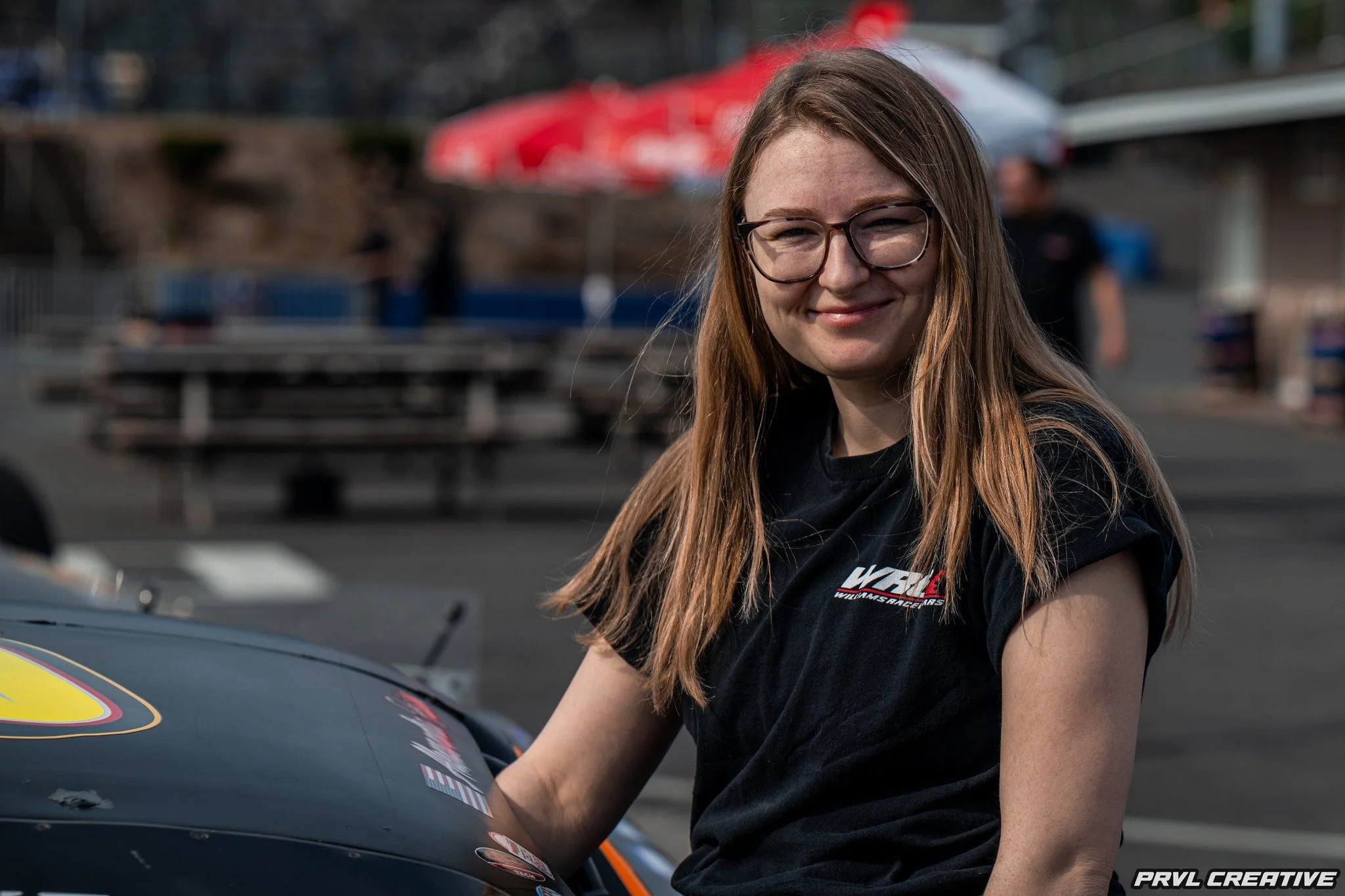 A young woman with long red hair and glasses smiling outdoors in front of a race car, wearing a black T-shirt with a logo.