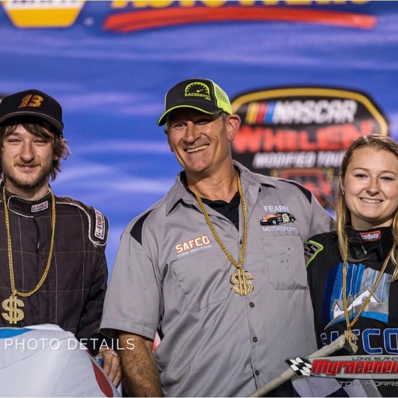 Three race car drivers, two men and one woman, wearing racing suits and gold dollar sign necklaces, standing in front of a NASCAR banner, smiling after a race event.