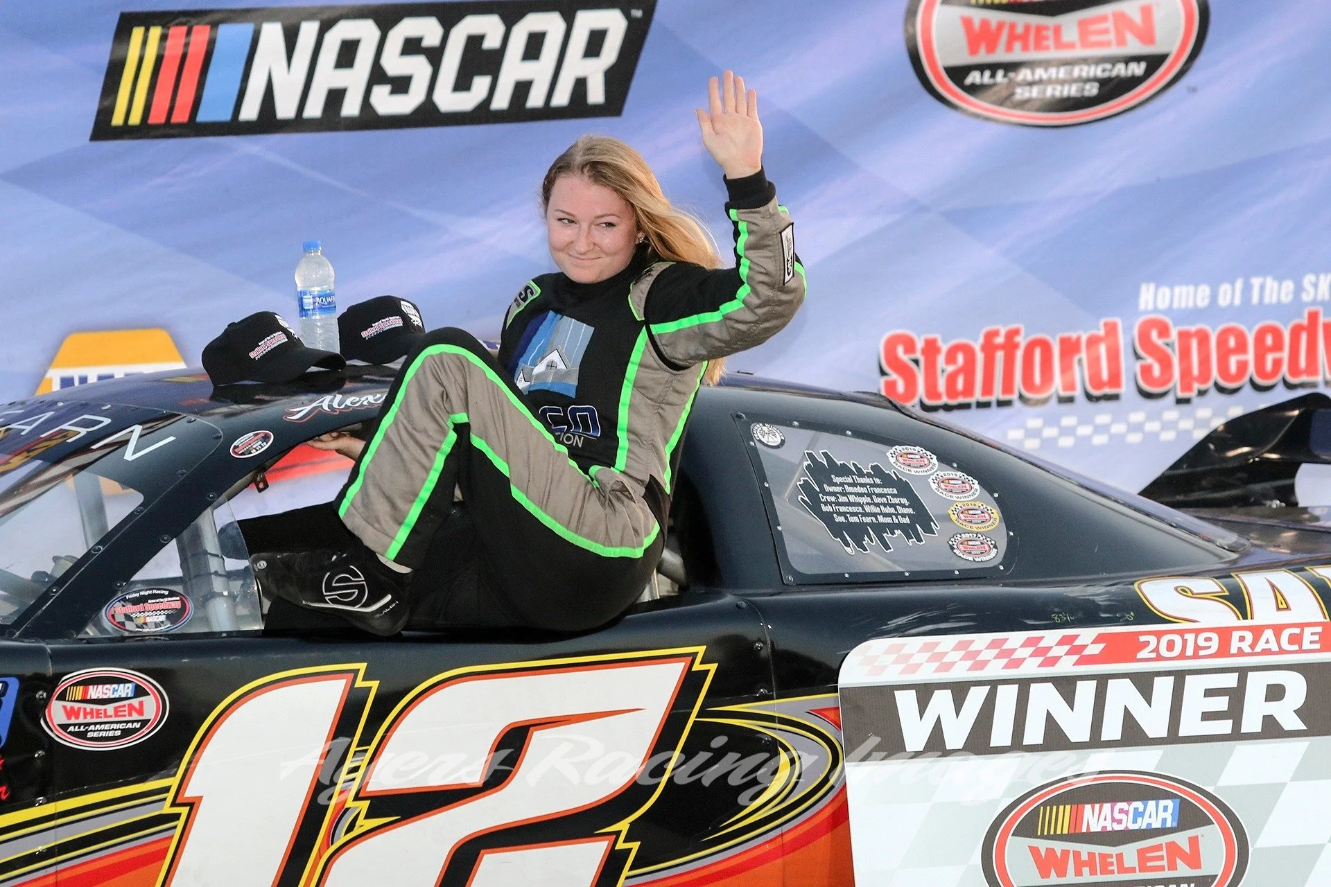 A woman in a racing suit sitting on top of a race car, waving and smiling, in front of a backdrop with NASCAR and Stafford Speedway logos, indicating she is the winner of a 2019 race.