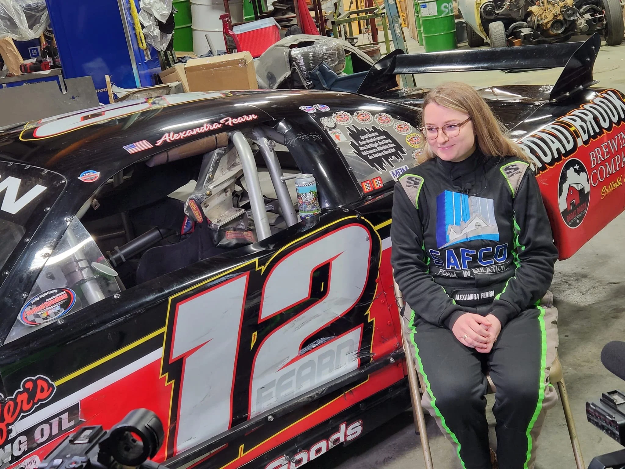 A woman in racing gear sitting on a chair next to a black and red race car in a garage.