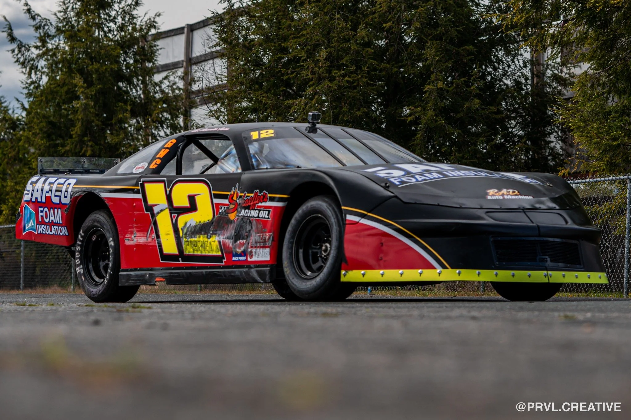 Black and red race car with the number 12, sponsor logos including Safeco Foam Insulation, and a yellow front lip, parked on an asphalt surface with trees and a fence in the background.
