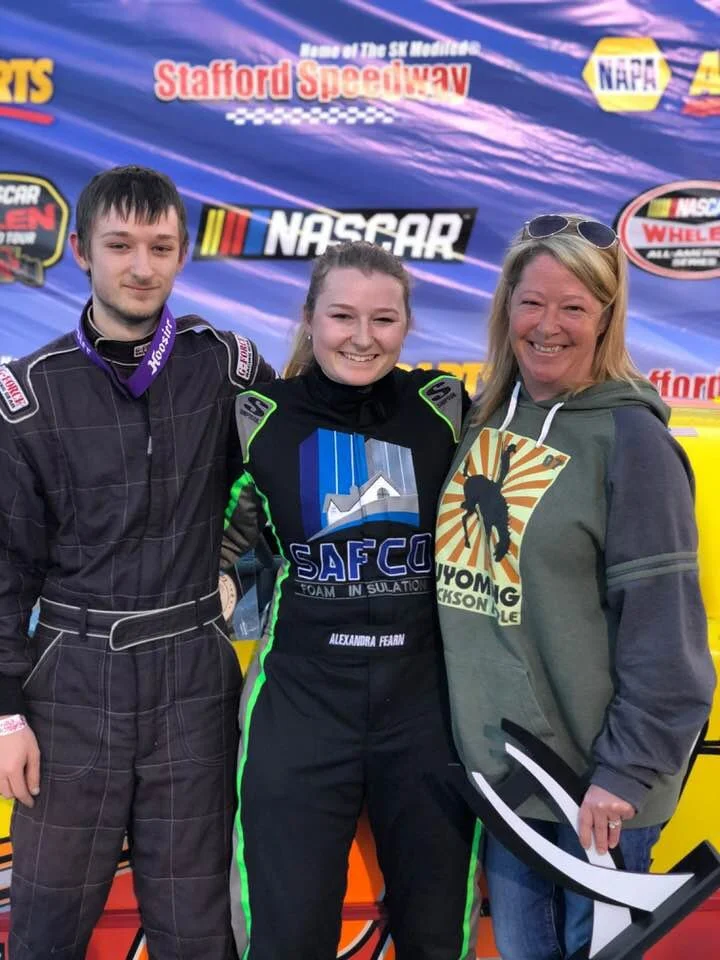 Three people standing together at a racing event, with a backdrop displaying NASCAR, Stafford Speedway, and other racing sponsors. The person in the middle is a race car driver wearing a black and green racing suit with the logo "SAFCO". The person o