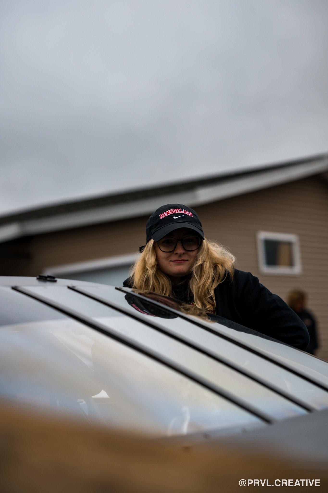 A woman with blond hair, glasses, and a black cap stands behind a metallic car, looking at the camera under a cloudy sky, with a house in the background.