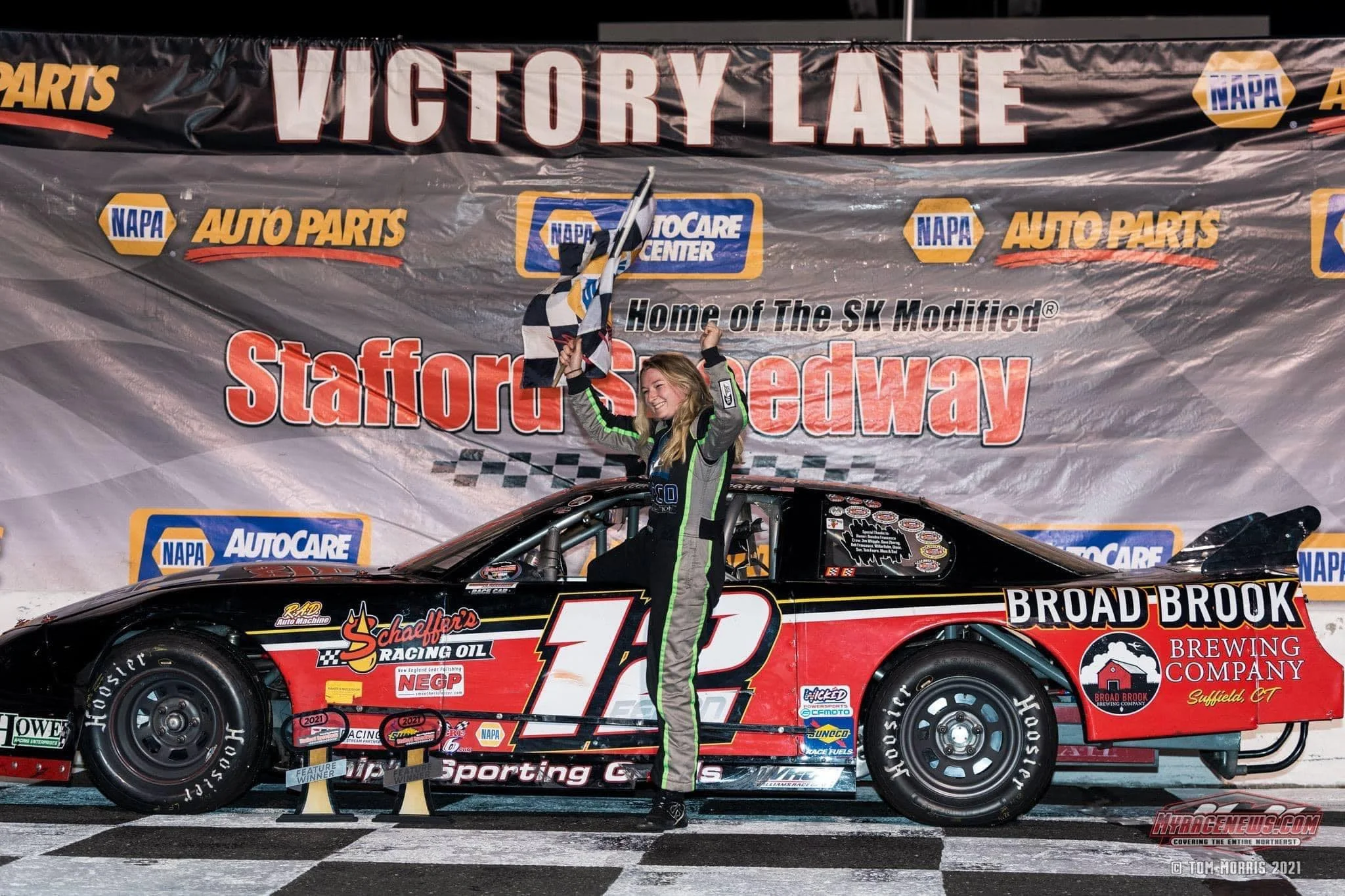 A young female race car driver celebrates victory on the winner's podium at Stafford Motor Speedway, holding a checkered flag and standing next to her black and red race car with the number 12. She wears a racing suit and is smiling with one arm rais