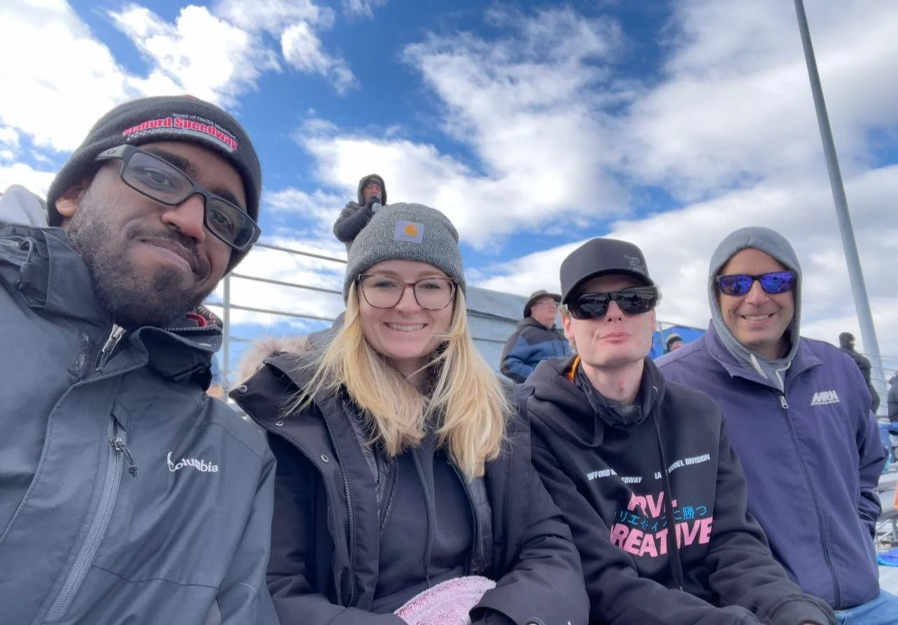 Group of four people sitting outdoors on bleachers at a sporting event, dressed in winter clothing, with a clear blue sky and clouds overhead.