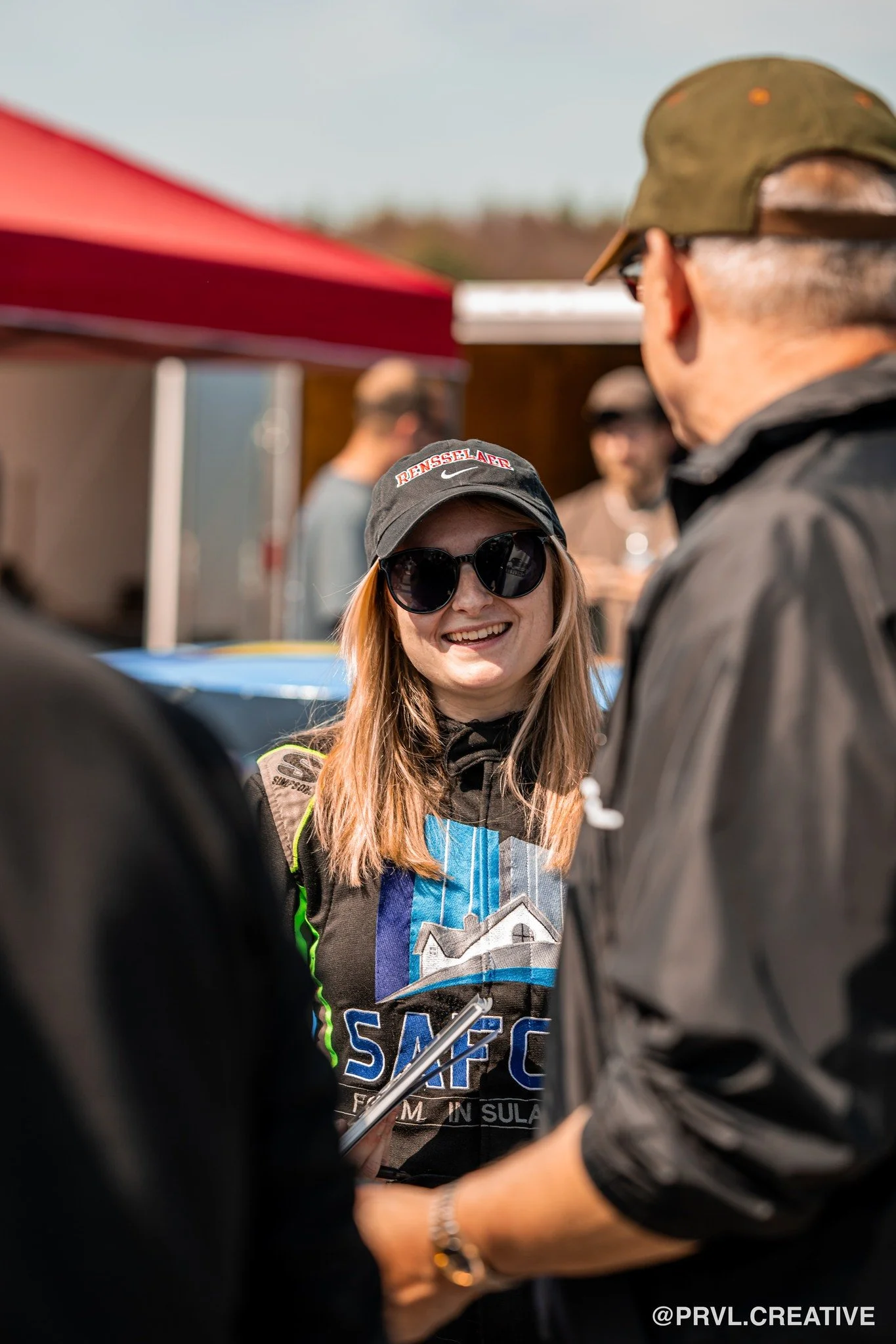 A young woman smiling and wearing sunglasses, a cap, and a racing suit, talking to a man at an outdoor event. Other people and tents are visible in the background.