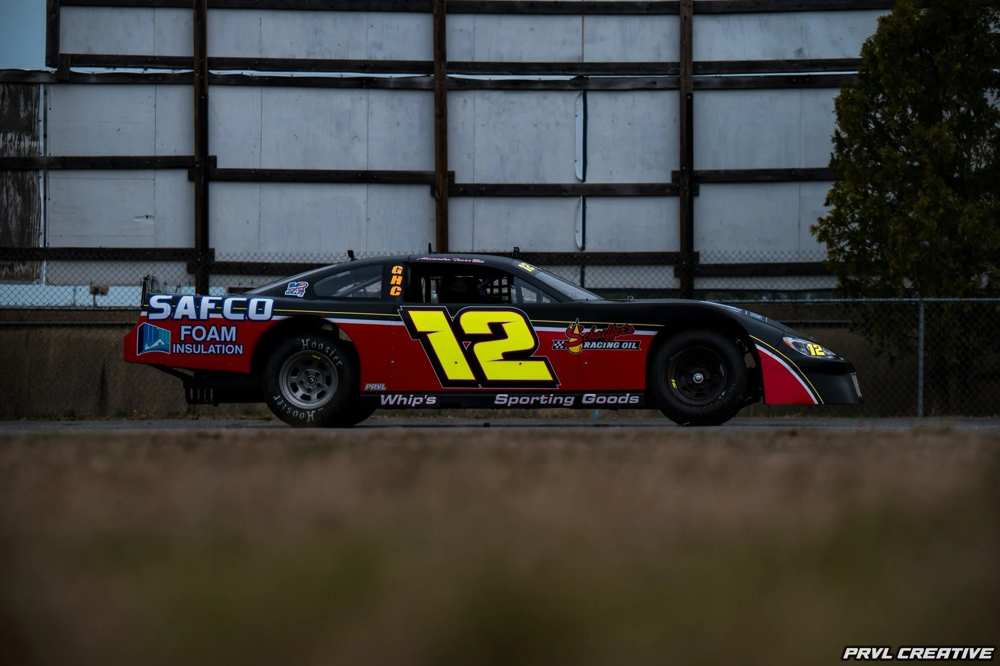 Race car with the number 12 on its side parked on a track, featuring branding for SAFCO Foam Insulation, Schafer's Racing Oil, and sporting a black, red, and yellow color scheme, with a background of a fence and a building.