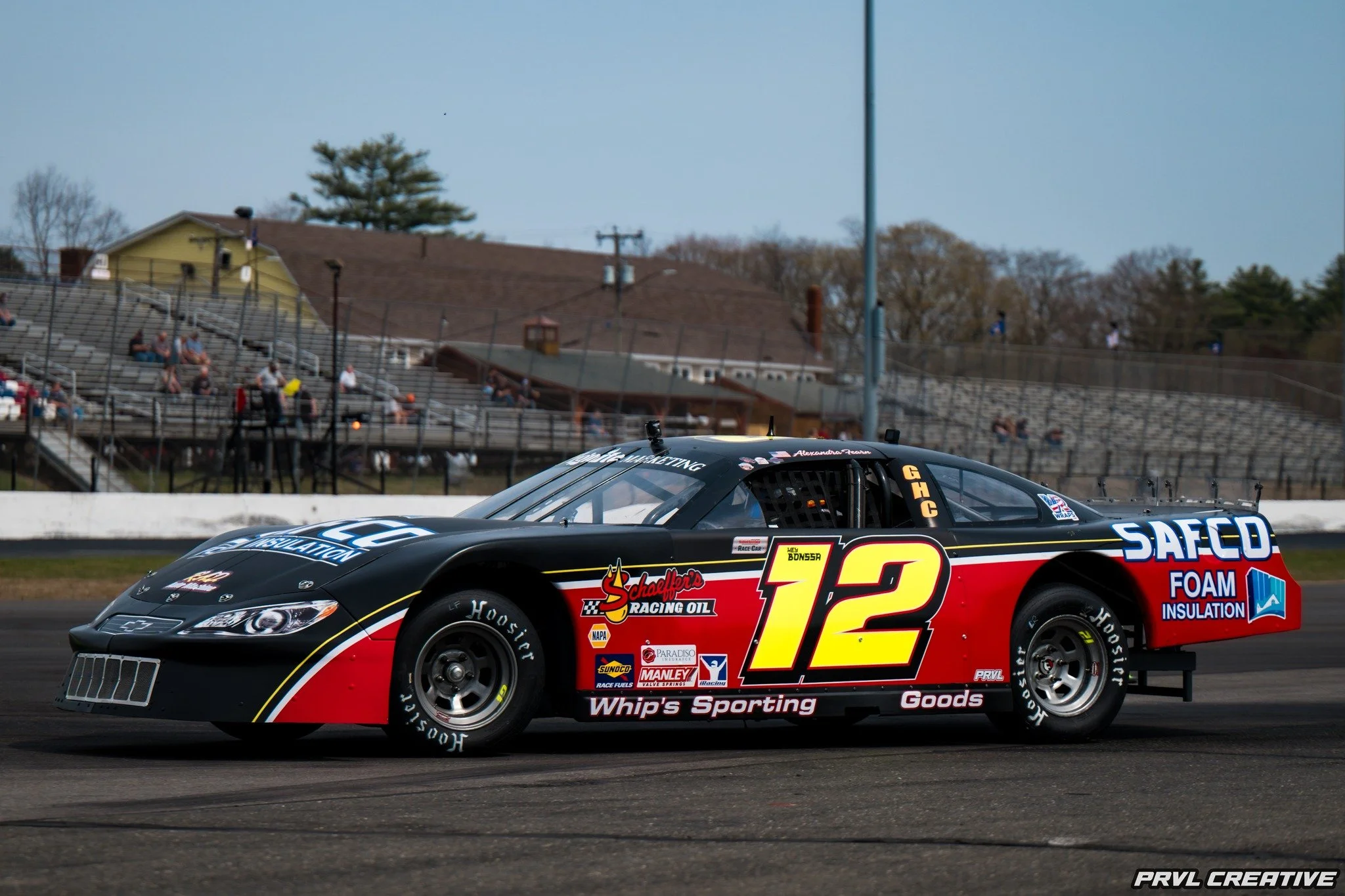 Black and red race car with number 12 on the side, racing on a track with spectators in bleachers in the background.