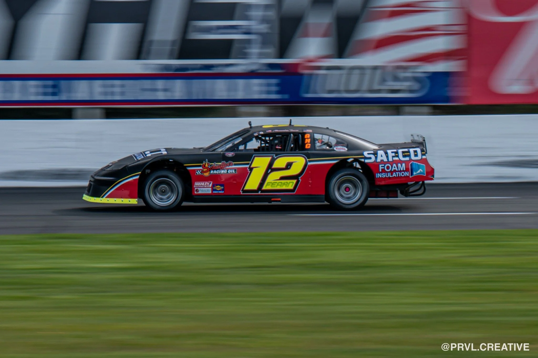 Race car number 12 on a racing track with blurred background and foreground indicating motion, featuring red, black, and yellow colors with various sponsor logos.